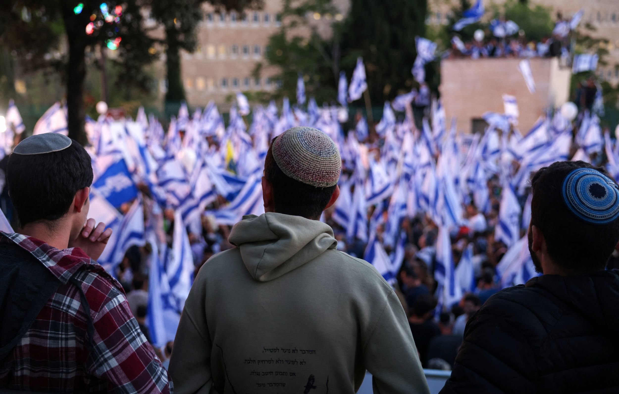 The image shows three individuals, viewed from behind, observing a large crowd at an outdoor event. The crowd is holding blue and white flags, which may symbolize a national identity. The individuals are wearing kippahs (traditional Jewish head coverings). The setting appears to be a public gathering, possibly a rally or demonstration, with trees and buildings visible in the background. The atmosphere seems lively and engaged.