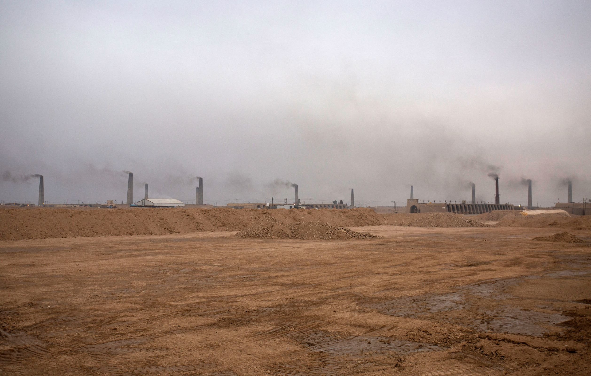 L'image montre un paysage industriel avec un sol désertique et nu. À l'arrière-plan, de nombreux cheminées émettent de la fumée dans une atmosphère brumeuse et chargée. Le ciel est grisâtre, ce qui donne une impression de pollution et de dégradation environnementale. Le terrain est principalement constitué de sable ou de terre, et l'ensemble dégage une atmosphère morose et industrialisée.
