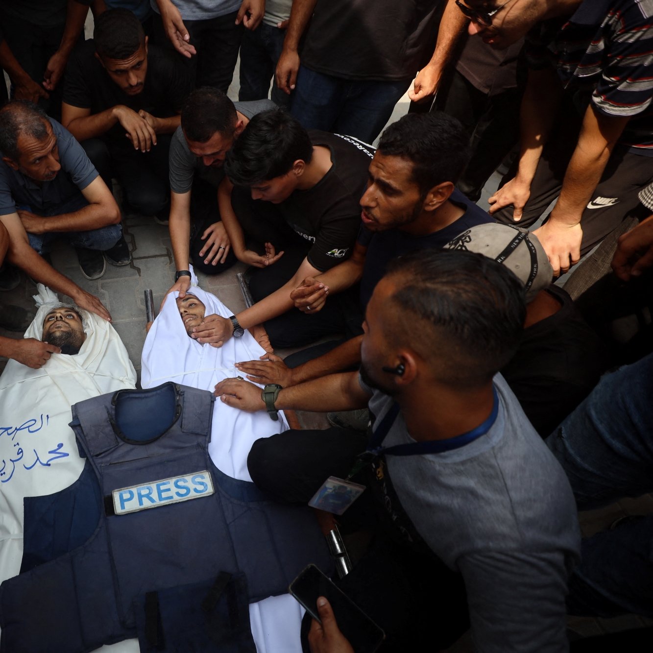 تصویری از گروهی از مردان که دور یک فرد دراز کشیده با پوششی به رنگ سفید و لباس خبرنگاری جمع شده‌اند.