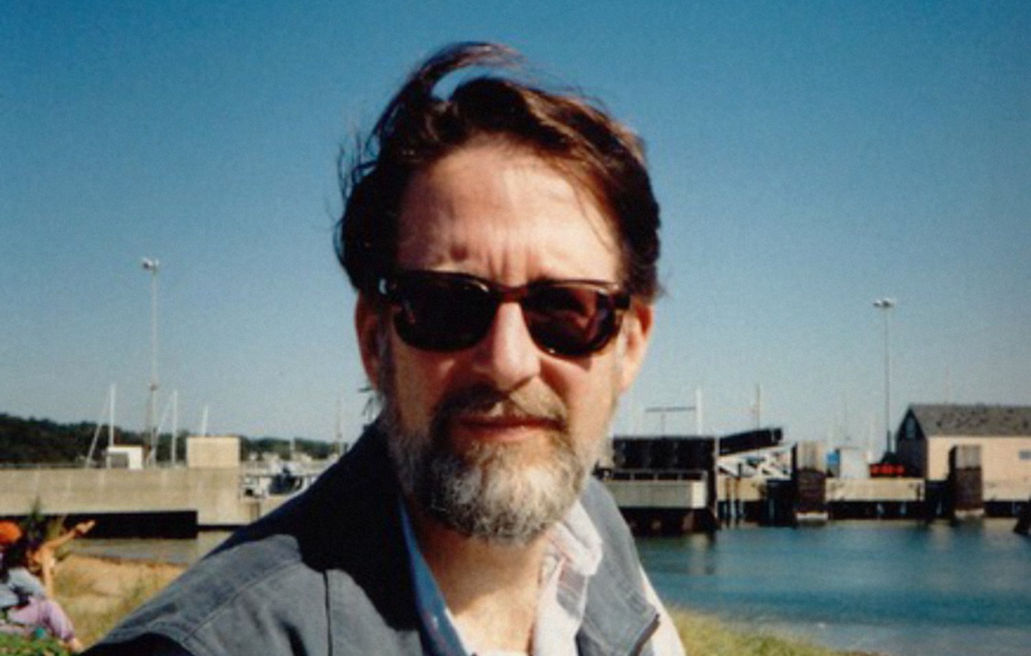 Je ne sais pas qui est cette personne, mais je peux décrire l'image. On y voit un homme avec des lunettes de soleil et une barbe, assis près de l'eau. Il a les cheveux bruns et un look décontracté, portant une veste. En arrière-plan, on aperçoit des bateaux et un quai, ce qui donne une ambiance maritime. Le ciel est clair et ensoleillé.