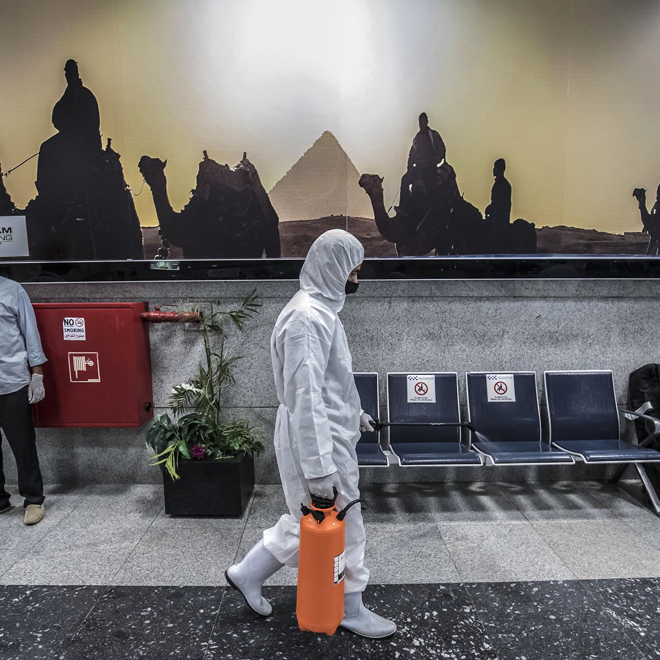 L'image montre un hall d'aéroport ou une station, avec un fond représentant des silhouettes de chameaux et de pyramides, visiblement au coucher du soleil. Au premier plan, une personne en costume de protection blanc marche, tenant une valise orange. Deux personnes en uniforme, probablement du personnel de sécurité ou de santé, se tiennent sur le côté, portant des masques. L'environnement est moderne et propre, avec des sièges en métal et des éléments de décoration comme des plantes.