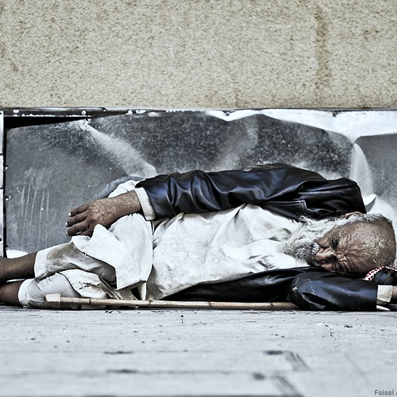 L'image montre un homme âgé allongé sur le sol, semblant endormi. Il porte des vêtements usés et semble être dans une position de repos sur un trottoir. Son visage est marqué par le temps et il est entouré d'un environnement urbain. L'atmosphère évoque une certaine mélancolie et une réflexion sur la vie des personnes sans abri.
