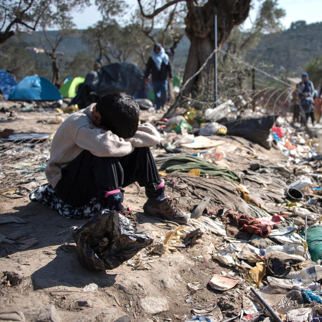 L'image montre un jeune enfant assis au sol, semblant triste ou désemparé. Il est entouré de déchets et de débris, ce qui souligne une situation de pauvreté et de désespoir. À l'arrière-plan, on peut voir d'autres personnes marchant, probablement des familles, dans un environnement difficile. Des tentes sont visibles, indiquant un campement temporaire. L'ensemble de la scène évoque une réalité difficile et poignante.
