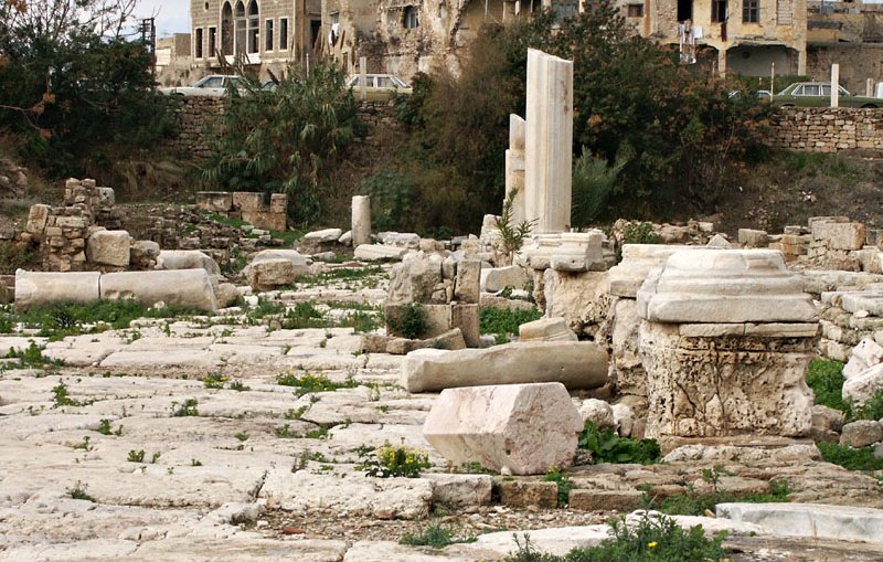 L'image montre un site archéologique avec des ruines anciennes. On peut voir des colonnes et des blocs de pierre, certains sont en désordre, tandis que d'autres semblent être en place. En arrière-plan, il y a des bâtiments plus récents, probablement des habitations, qui contrastent avec les vestiges historiques. La végétation est présente autour des pierres, avec des herbes et des fleurs qui poussent dans les fissures. L'ensemble évoque un mélange de l'histoire ancienne et de la vie moderne.