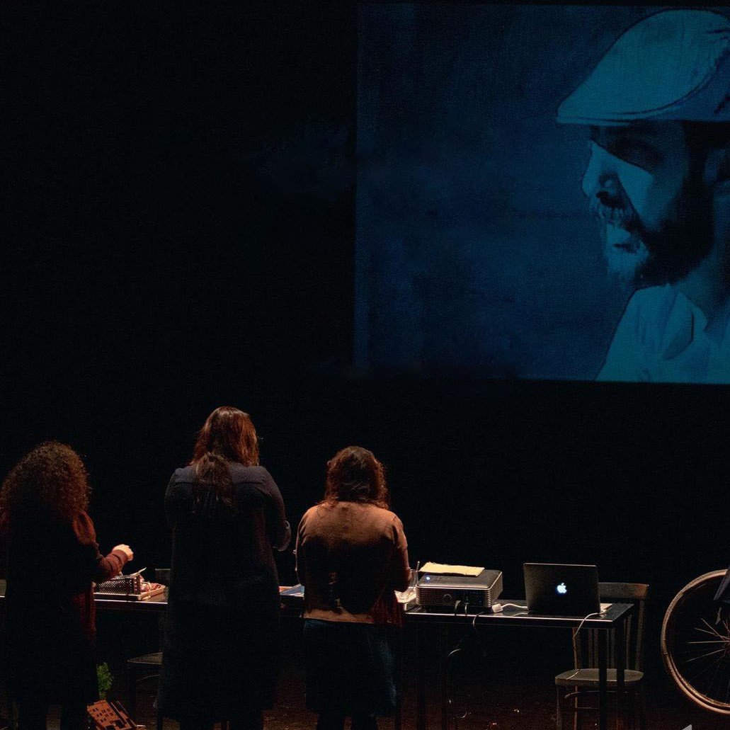 L'image montre un groupe de personnes de dos, se tenant devant une projection qui affiche une image en noir et blanc d'un homme portant un chapeau. Les personnes semblent engagées dans une activité, peut-être en lien avec la projection. Au premier plan, il y a un bureau avec un ordinateur et d'autres objets. L'éclairage crée une atmosphère intrigante autour de la scène.