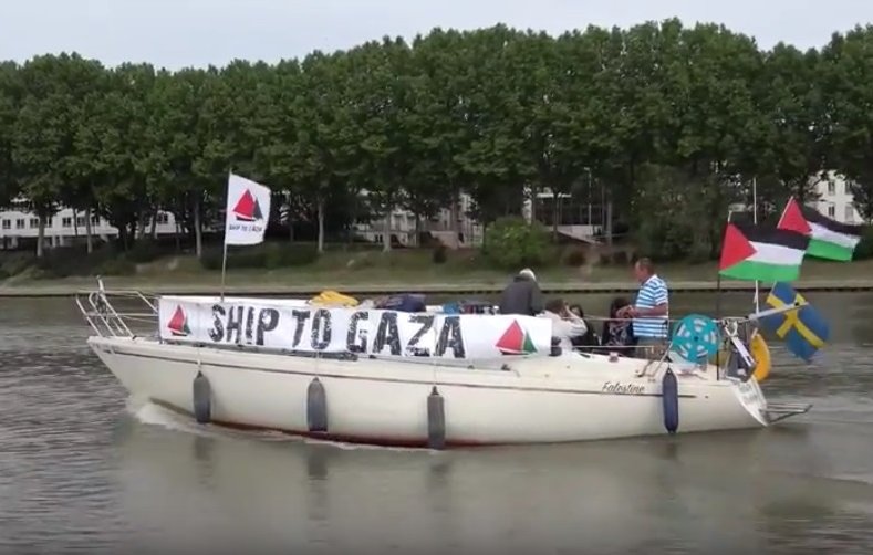 L'image montre un bateau naviguant sur une étendue d'eau. Sur le bateau, il y a des personnes présentes, et une banderole est accrochée, indiquant "SHIP TO GAZA". Le bateau arbore également plusieurs drapeaux, dont ceux de la Palestine et de la Suède, ainsi que d'autres drapeaux qui pourraient représenter différentes organisations ou pays. En arrière-plan, des arbres et des bâtiments sont visibles, suggérant un environnement urbain proche d'une rivière ou d'un port.