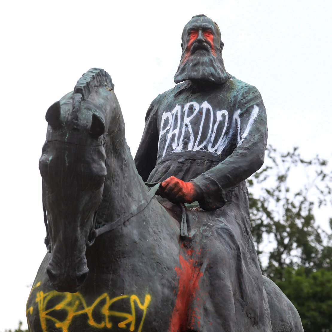 L'image montre une statue équestre d'un homme, probablement un personnage historique, qui porte des graffitis. Le mot "PARDON" est écrit en lettres blanches sur sa robe, tandis que d'autres mots, comme "RACIST", apparaissent en couleur sur le socle. La statue semble être recouverte de peinture, ce qui suggère un acte de protestation ou de critique. En arrière-plan, on peut voir un ciel nuageux et quelques arbres.
