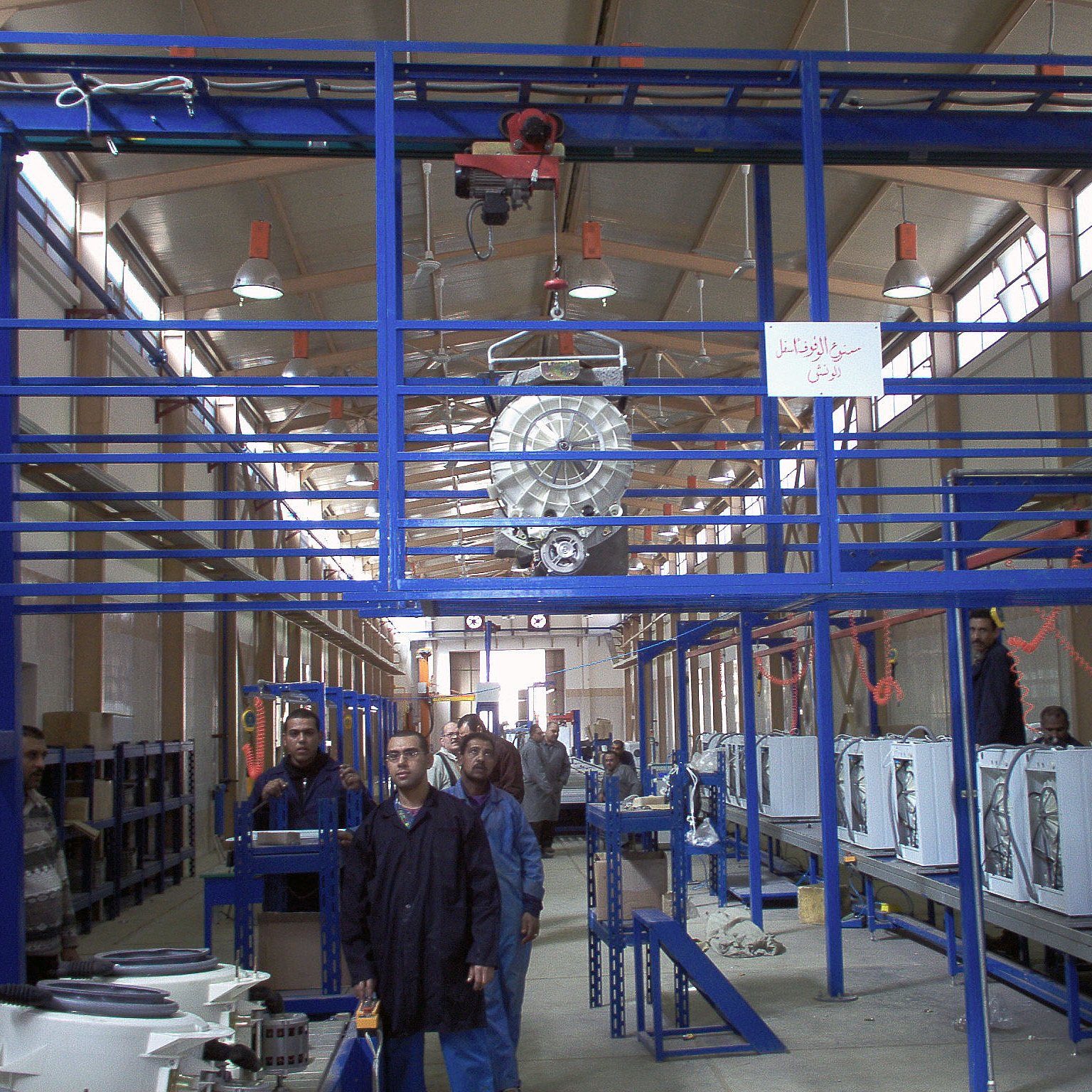 The image shows an industrial setting, likely a manufacturing or assembly facility. There are several workers in the foreground, wearing uniforms, and they seem to be engaged in tasks related to assembling or inspecting products. Above them, there is overhead equipment, possibly a crane or hoist, used for lifting materials. The facility has rows of workstations with what appears to be appliances or machinery on display, indicating a focus on production. The interior is well-lit with a spacious design, featuring high ceilings and a structured layout.