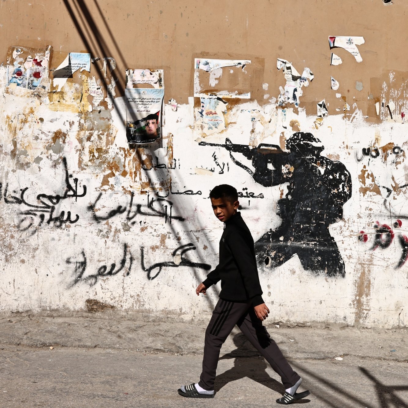 A boy walks past a wall with a soldier graffiti and Arabic text, contrasting street life.