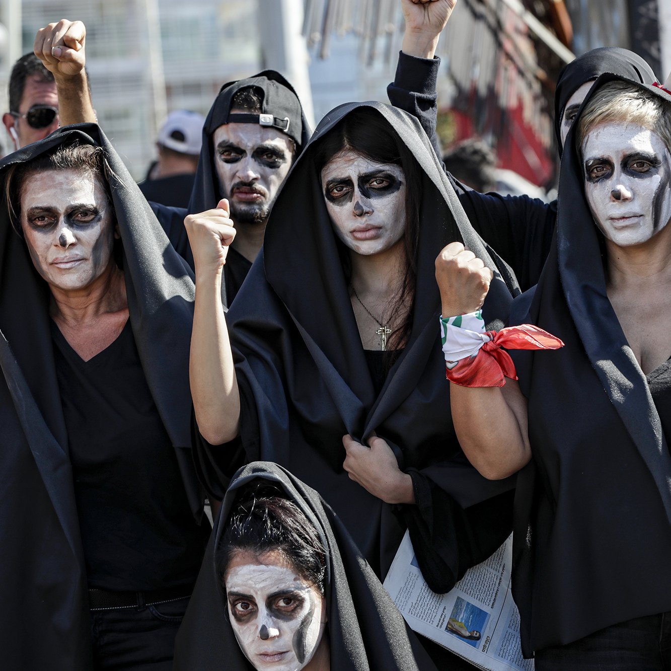 The image depicts a group of people wearing black hooded cloaks and painted faces, featuring white and dark makeup that gives a striking visual effect. They appear to be raising their fists in a gesture of solidarity or protest. The atmosphere suggests they are part of a demonstration or performance, possibly conveying a message or advocating for a specific cause. The backdrop includes hints of a larger gathering, emphasizing the collective nature of their presence.