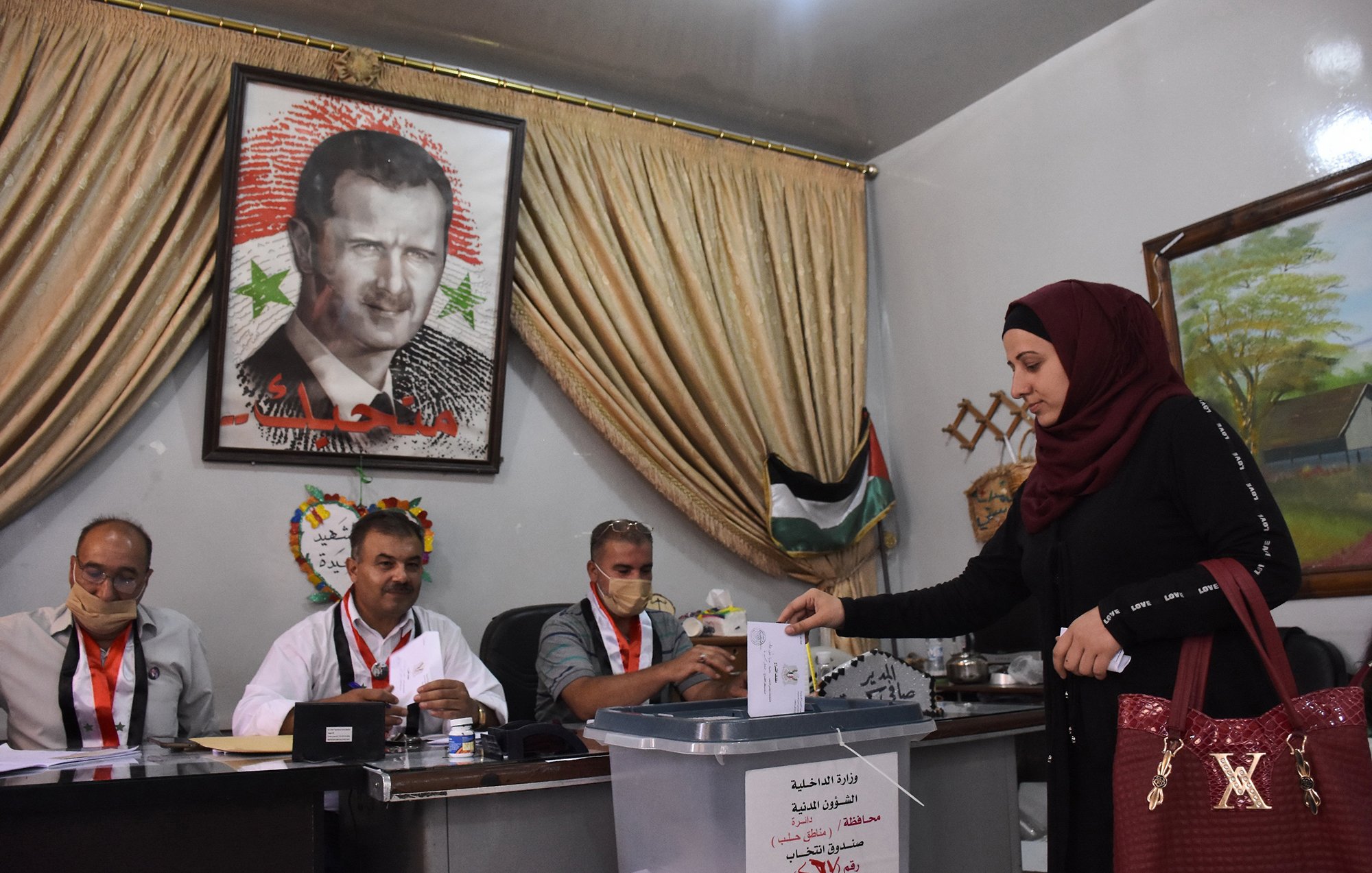 L'image montre une scène de vote, probablement lors d'élections en Syrie. Au premier plan, une femme en hijab dépose un bulletin dans une urne transparente. À l'arrière-plan, plusieurs hommes sont assis derrière une table, regardant le processus. Un grand portrait d'un homme, probablement un dirigeant, est accroché au mur, entouré de drapeaux et de décorations. Les participants portent des badges d'identification, et l'atmosphère semble sérieuse et formelle.