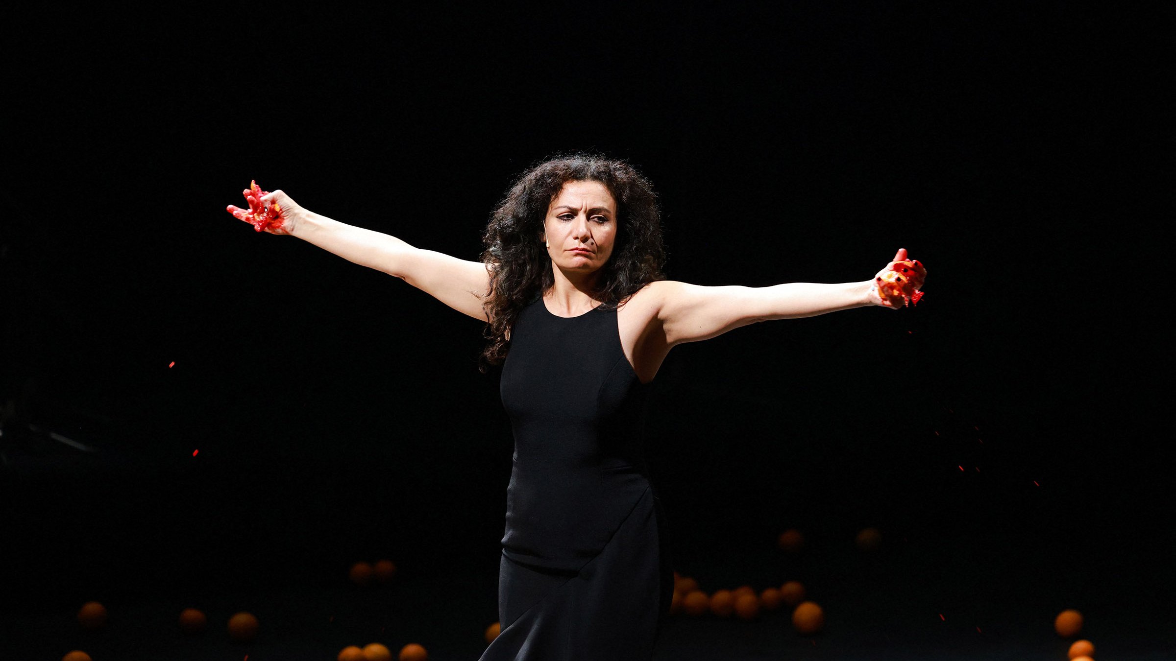 L'image montre une femme debout sur scène, les bras étendus de chaque côté. Elle porte une robe noire et exprime une émotion intense. Des oranges sont dispersées autour d'elle sur le sol, ajoutant une dimension visuelle frappante à la scène. Des éclaboussures de liquide rouge semblent provenir de ses mains, ce qui accentue le drame et la tension de l'instant. L'éclairage de la scène est sombre, mettant ainsi en valeur la silhouette de la femme et les oranges.