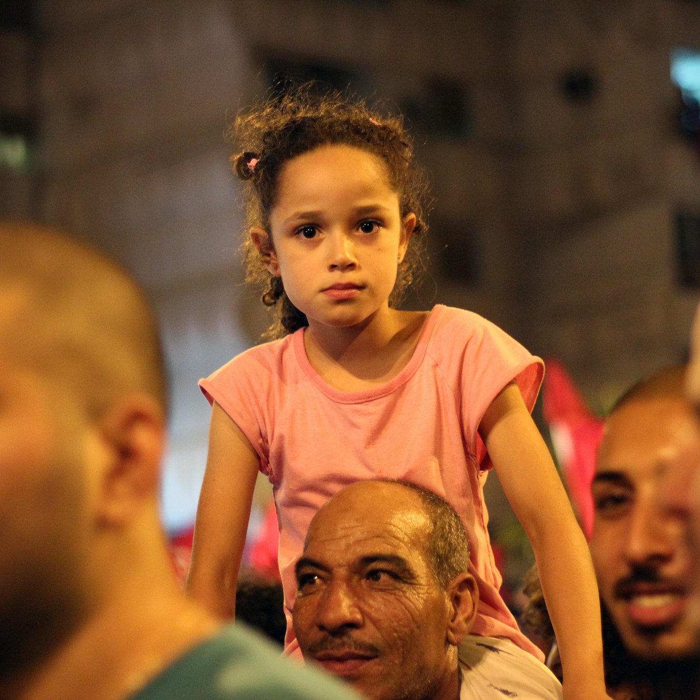 L'image montre une petite fille avec des cheveux bouclés, assise sur les épaules d'un homme dans une foule. Elle porte un t-shirt rose et a une expression sérieuse. Autour d'elle, on peut voir des visages d'adultes, certains souriant et d'autres concentrés, ce qui suggère une ambiance dynamique, peut-être lors d'un événement ou d'une célébration. L'arrière-plan est flou, mais on peut distinguer des lumières et des drapeaux, ajoutant à l'atmosphère animée de la scène.