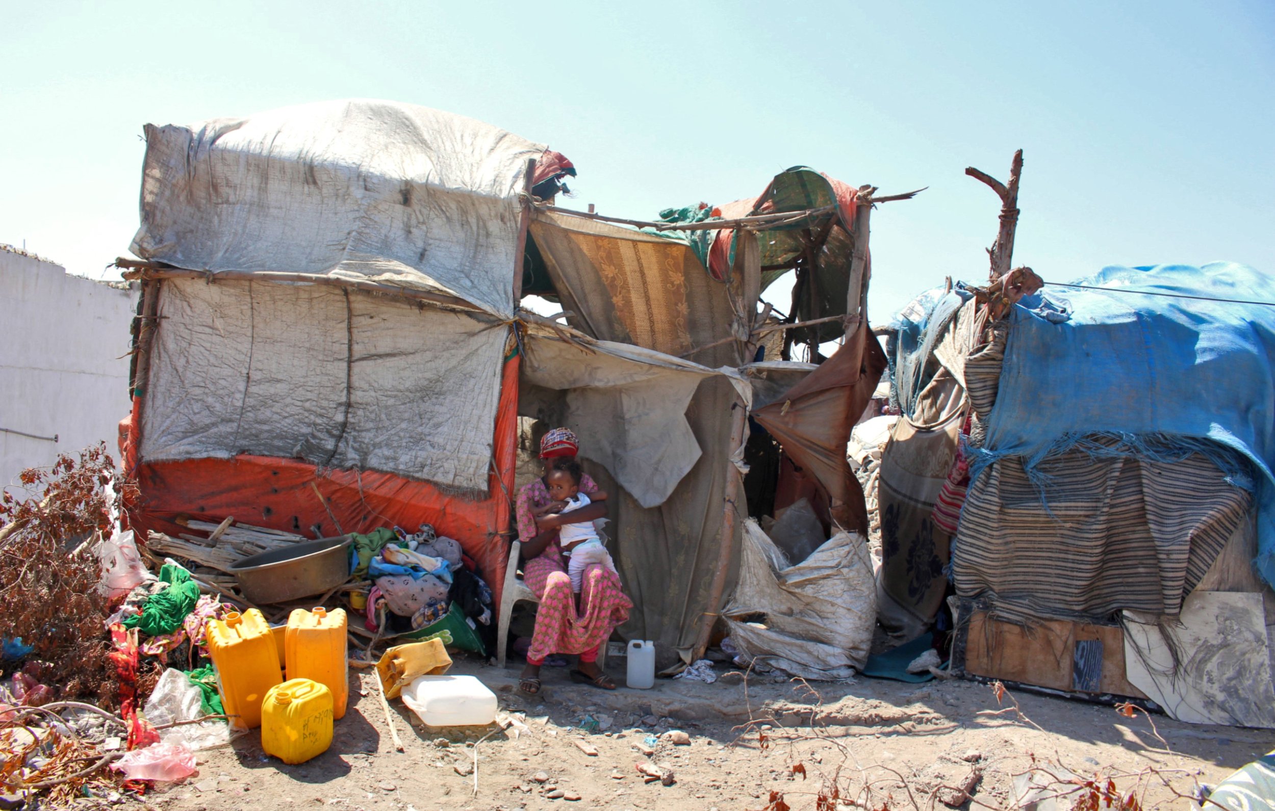 L'image montre une habitation informelle construite avec des matériaux comme des bâches en plastique et des morceaux de toile. À l'avant, on peut voir une femme assise dans l'ombre de la structure, entourée de divers objets et contenants, probablement utilisés pour la vie quotidienne. L'environnement semble un peu désordonné, avec des débris et des matériaux épars autour de l'habitation. Le ciel est dégagé, ce qui donne une impression de chaleur.
