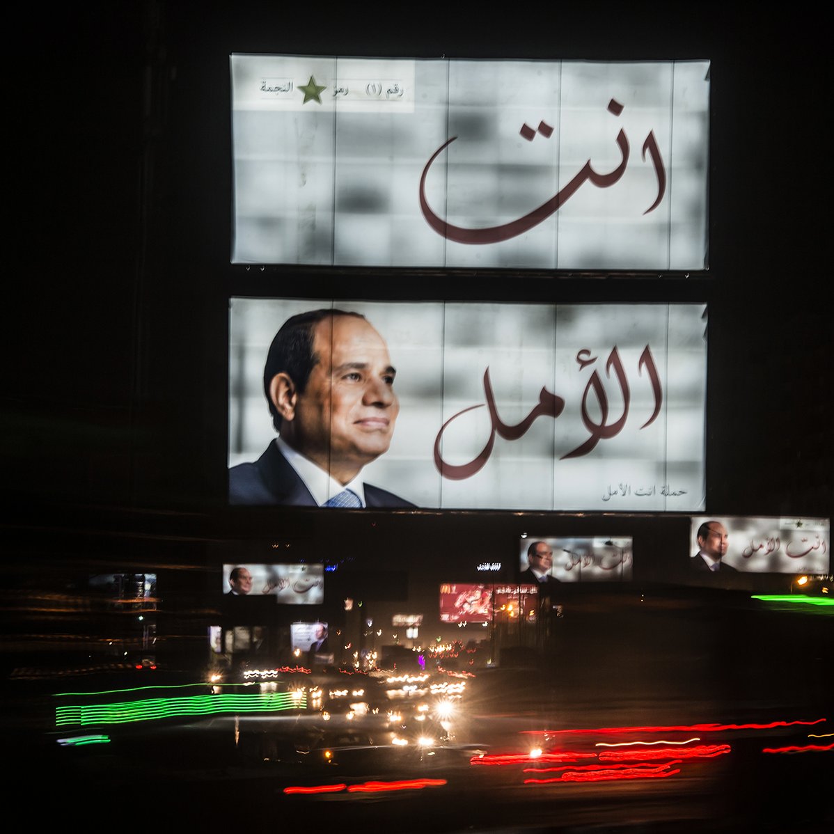 The image shows large billboards prominently displaying Arabic text. One of the billboards features a portrait of a man, likely a political figure, along with the words "انت الأمل," which translates to "You are the hope." The setting appears to be a busy urban area at night, with car lights creating a dynamic effect, suggesting movement and activity. The overall atmosphere reflects a lively city scene with political messaging.