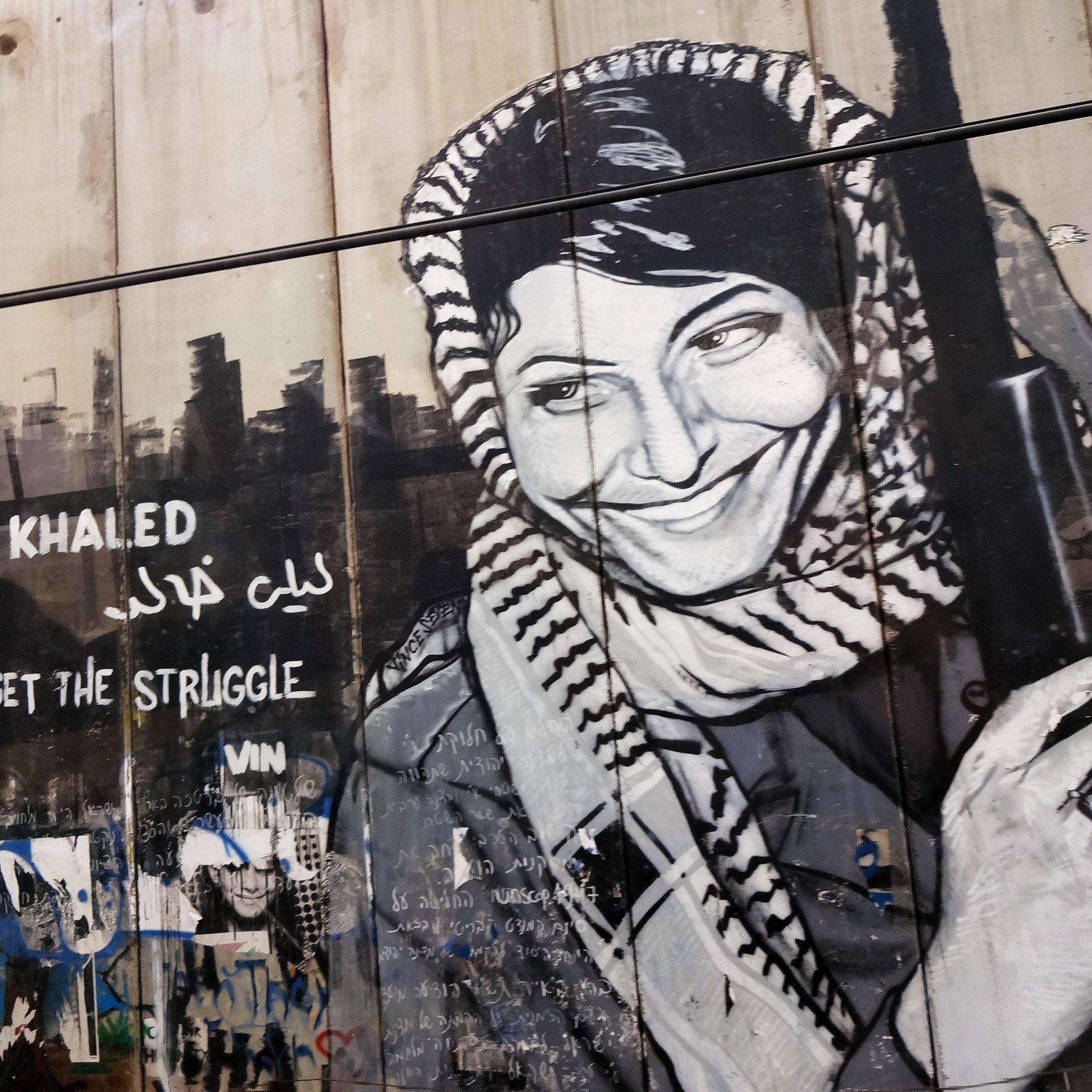 L'image présente une fresque murale représentant Leïla Khaled, une figure emblématique de la résistance palestinienne. Elle est dépeinte avec un sourire, portant une écharpe typique et tenant un fusil. À côté de son portrait, on peut lire des inscriptions en anglais et en arabe, telles que "Don't forget the struggle" et son nom. L'arrière-plan est constitué d'un mur, accentuant le message de lutte et de mémoire associé à son image. Des graffitis colorés et des marques diverses décorent également la surface, ajoutant à l'atmosphère militante et artistique du lieu.