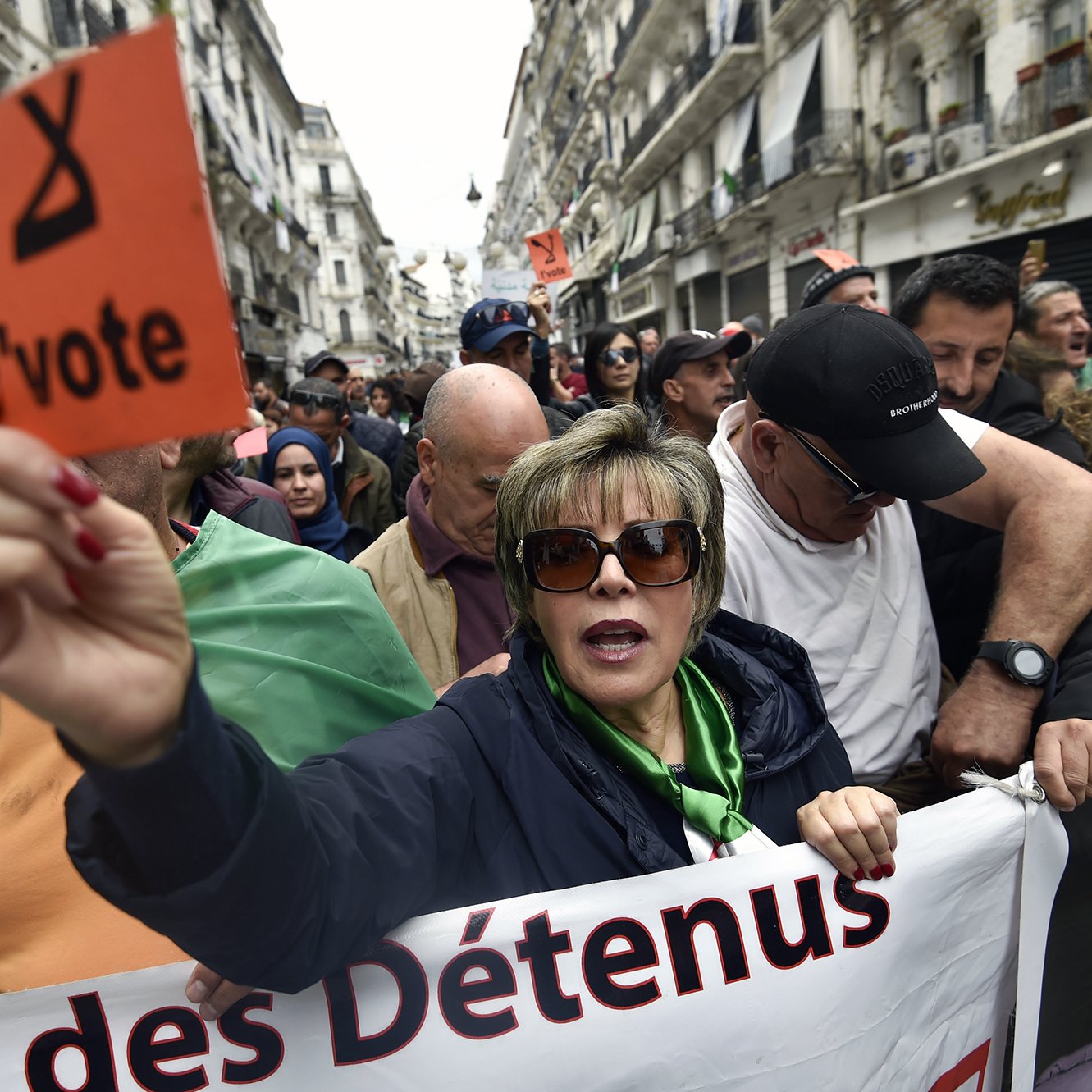 L'image montre une manifestation dans une rue d'Alger. Au premier plan, une femme tient une pancarte orange avec le mot "l'vote". Elle est entourée par d'autres manifestants, dont certains portent des drapeaux algériens. L'atmosphère semble engagée et déterminée, avec des expressions de solidarité parmi les participants. Des banderoles sont visibles, indiquant des revendications liées à la situation politique. L'environnement urbain et les bâtiments en arrière-plan témoignent d'une ambiance dynamique de protestation.