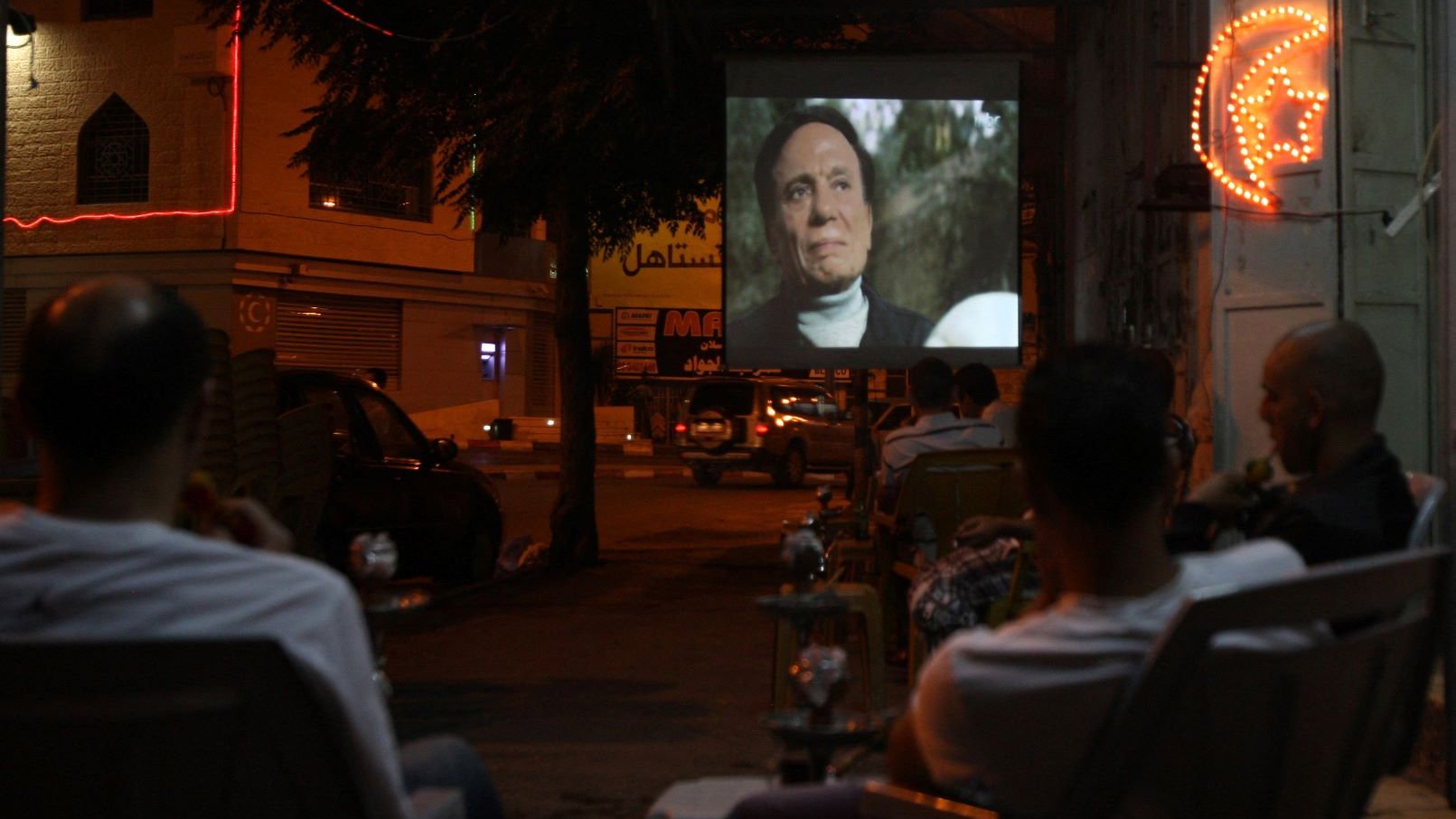 L'image montre une scène nocturne où un groupe de personnes est assis à l'extérieur, observant un film projeté sur un mur. On voit des chaises disposées face à l'écran, et quelques fumées de narguilé flottent dans l'air. L'environnement est animé par des lumières de néon, et on aperçoit des voitures garées et des bâtiments en arrière-plan, créant une ambiance conviviale et détendue.