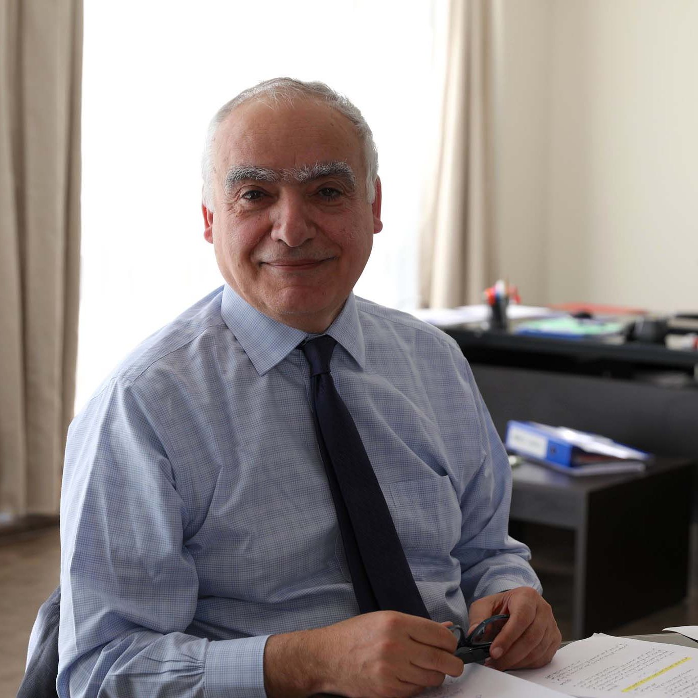 Je ne peux pas identifier les personnes dans l'image. Cependant, je peux décrire la scène : on voit un homme souriant assis à un bureau, avec des papiers devant lui. La pièce semble bien éclairée, avec une lumière naturelle. En arrière-plan, on aperçoit des dossiers et un meuble de rangement. L'homme porte une chemise et une cravate.