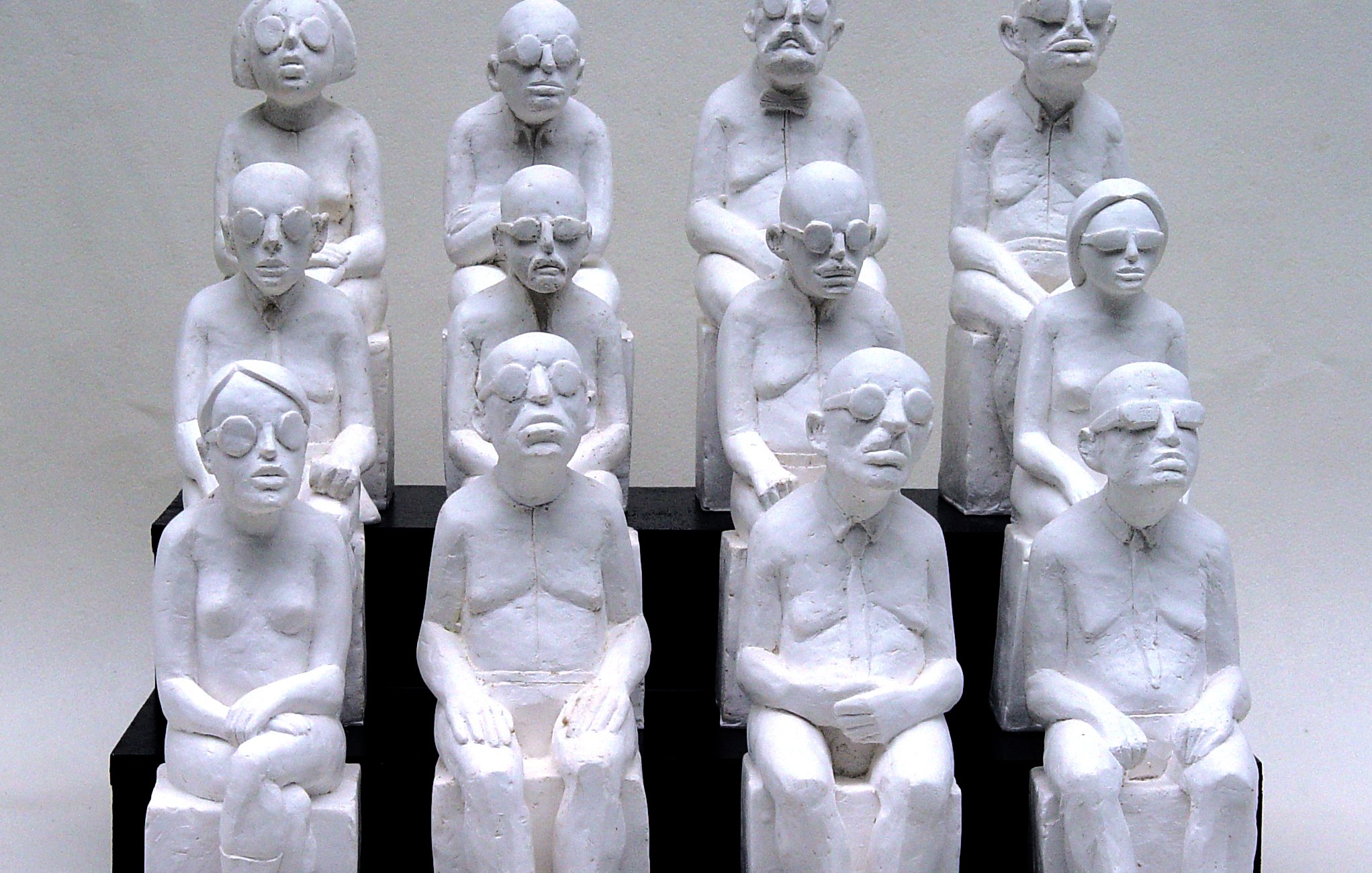 L'image représente une sculpture où plusieurs figures humaines, toutes de couleur blanche, sont assises sur des bancs. Les personnages sont stylisés avec des traits simples et géométriques, portant des lunettes, et semblent avoir des expressions neutres ou pensive. Ils sont agencés en un groupe de dix, disposés en plusieurs rangées, créant une impression d'unité et de contemplation. Les sculptures semblent évoquer des thèmes tels que la solitude, la réflexion ou la condition humaine.