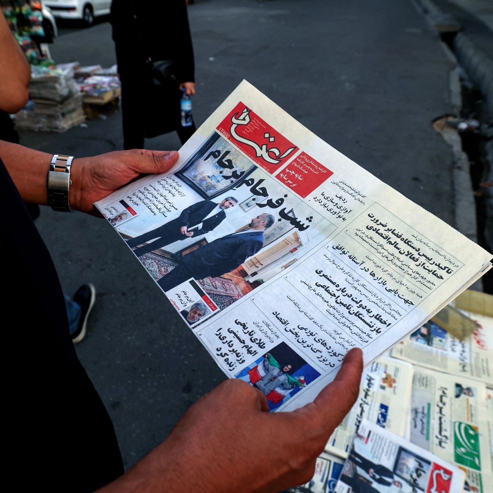 L'image montre une personne tenant un journal ouvert. Le titre est bien visible et semble relater des informations d'actualité, potentiellement politiques ou sociales. En arrière-plan, on aperçoit un kiosque à journaux avec d'autres exemplaires de journaux exposés. L'environnement semble urbain, avec des voitures garées et un sol en pavé.