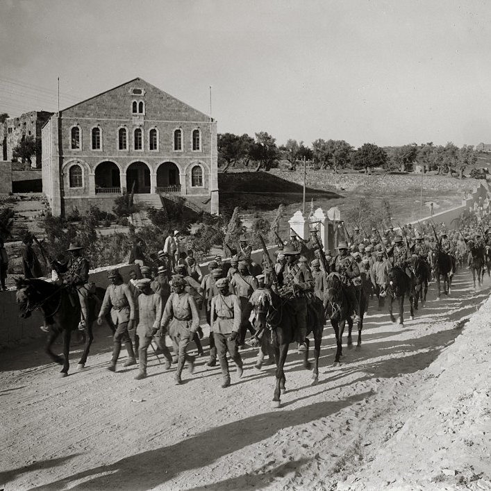 L'image montre une troupe de soldats à cheval marchant en formation le long d'un chemin. En arrière-plan, on peut voir des bâtiments en pierre, dont une grande structure avec des fenêtres. Le sol est poussiéreux, et on remarque également quelques personnes à pied, dont une femme en robe. L'atmosphère semble être celle d'une époque historique, probablement d'un conflit ou d'une quelconque mobilisation militaire. Les soldats portent des uniformes similaires et semblent déterminés dans leur avancée.