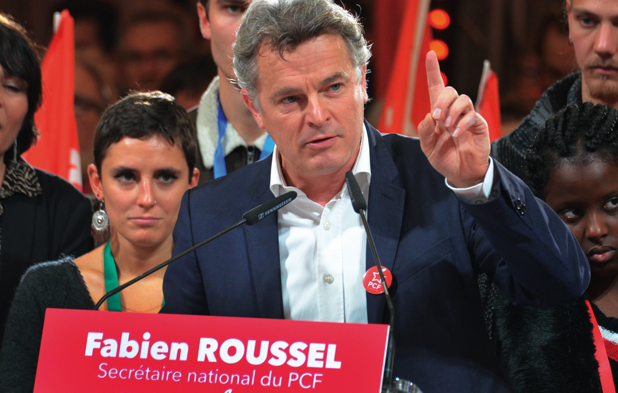 L'image montre un homme au premier plan, probablement en train de faire un discours. Il porte une veste et a l'air déterminé, levant un doigt comme s'il soulignait un point important. Derrière lui, plusieurs personnes l'écoutent, certaines regardant directement l'objectif. En bas de l'image, on peut voir une pancarte rouge avec le texte « Fabien ROUSSEL, Secrétaire national du PCF » et la date du 25 novembre 2018. L'ambiance semble être celle d'un congrès ou d'un rassemblement politique.