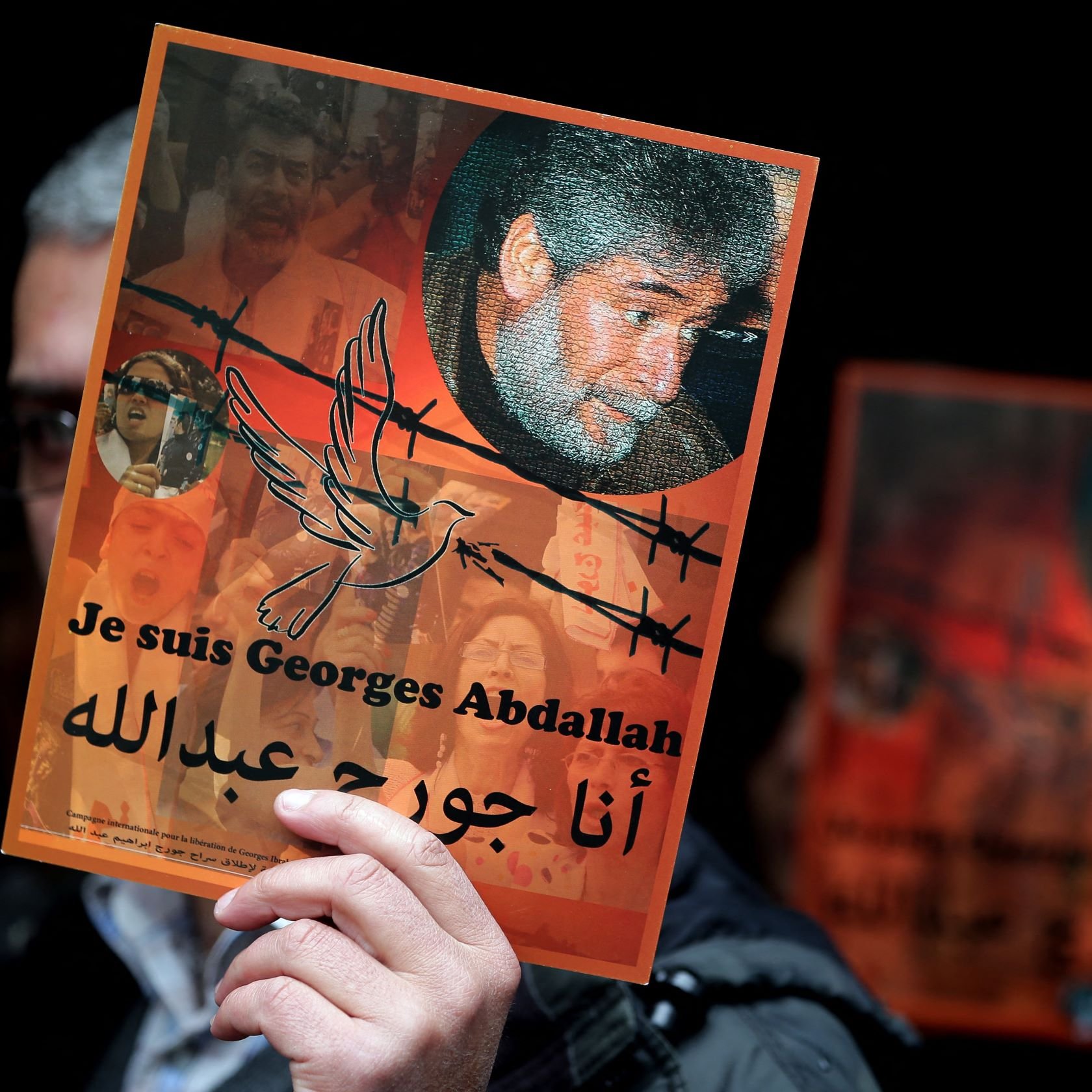 L'image montre une personne tenant une affiche. L'affiche met en avant le message "Je suis Georges Abdallah" en français et en arabe. Elle inclut un portrait de Georges Abdallah entouré de motifs symboliques, comme des barbelés et une colombe. En arrière-plan, on aperçoit des visages et des motifs qui suggèrent un contexte de soutien et d'engagement. L'ambiance semble être celle d'une manifestation ou d'un rassemblement pour la défense de ses droits.
