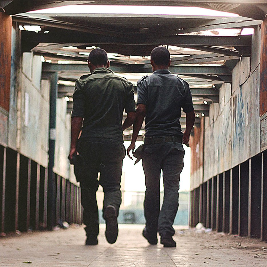 L'image montre deux hommes marchant côte à côte dans un passage couvert. Ils portent des uniformes de couleur sombre et se déplacent vers la sortie, avec leurs silhouettes se détachant contre la lumière. Les murs du passage semblent usés, avec des graffitis visibles, et le sol est pavé. L'atmosphère est à la fois urbaine et légèrement mélancolique, donnant une impression d'intimité entre les deux personnages.