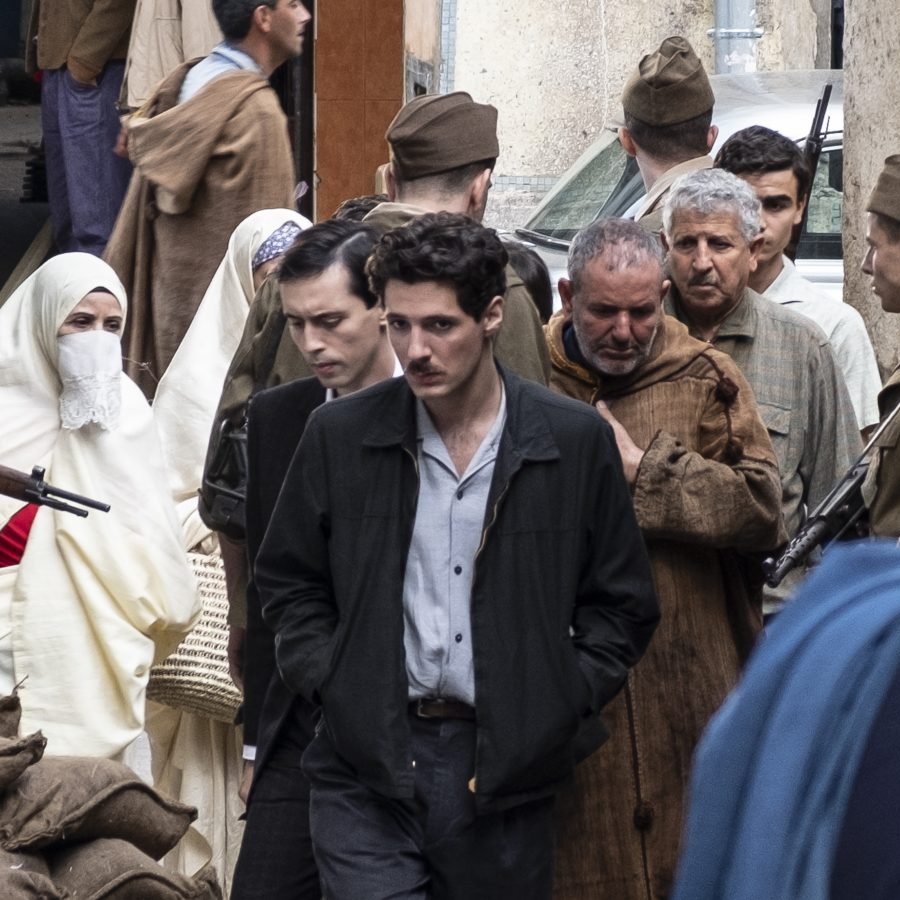 L'image montre une scène urbaine où plusieurs personnes sont rassemblées. Au centre, on voit un jeune homme en chemise claire et veste sombre, l'air pensif. Il est entouré de groupes de personnes, dont certaines semblent discuter ou se déplacer. Il y a également des hommes en uniformes militaires et des femmes portant des vêtements traditionnels, y compris un couvre-chef. L'ambiance de la scène semble chargée d'émotion, et le décor évoque une atmosphère historique ou dramatique.