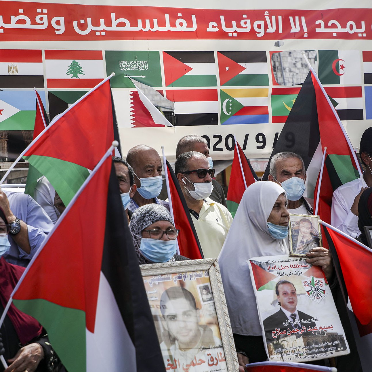L'image montre un groupe de personnes rassemblées, portant des masques et tenant des drapeaux. Ils semblent manifester pour soutenir la Palestine, avec des affiches et des portraits en mains. En arrière-plan, on peut voir une grande bannière ornée de multiples drapeaux de différents pays. L'ambiance est engagée et solidaire, témoignant d'une mobilisation pour une cause politique.
