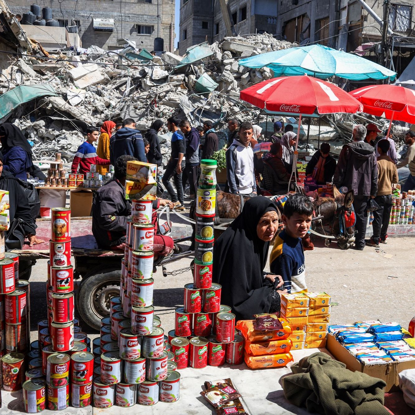 L'image montre une scène d'un marché en plein air, où des gens se déplacent parmi des étals de produits. Au premier plan, une femme en hijab et un enfant sont assis devant une pile de conserves et d'autres produits alimentaires. En arrière-plan, des bâtiments en ruines et des débris sont visibles, suggérant un contexte de destruction. Des parasols de différentes couleurs offrent de l'ombre aux vendeurs et aux clients. L'atmosphère semble chargée de vie malgré le décor dévasté.