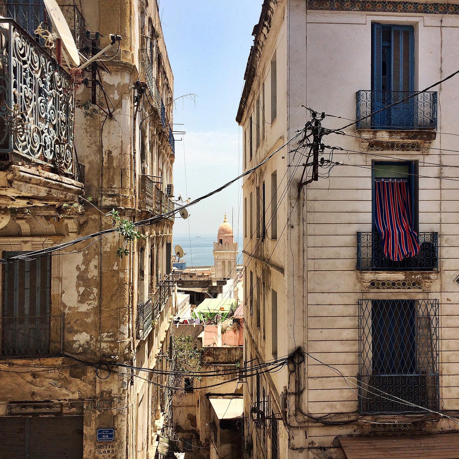 L'image montre une vue d'une rue étroite et animée d'une ville méditerranéenne. De chaque côté, on voit des bâtiments anciens avec des façades usées et des balcons en fer forgé. Des fils électriques serpentent entre les immeubles. Au loin, on peut apercevoir un dôme qui pourrait appartenir à une église ou une mosquée, et un bout de mer est visible, ajoutant une touche de bleu au paysage. Les fenêtres des bâtiments sont ornées de volets ou de rideaux, et l'ensemble dégage une atmosphère nostalgique et pittoresque.