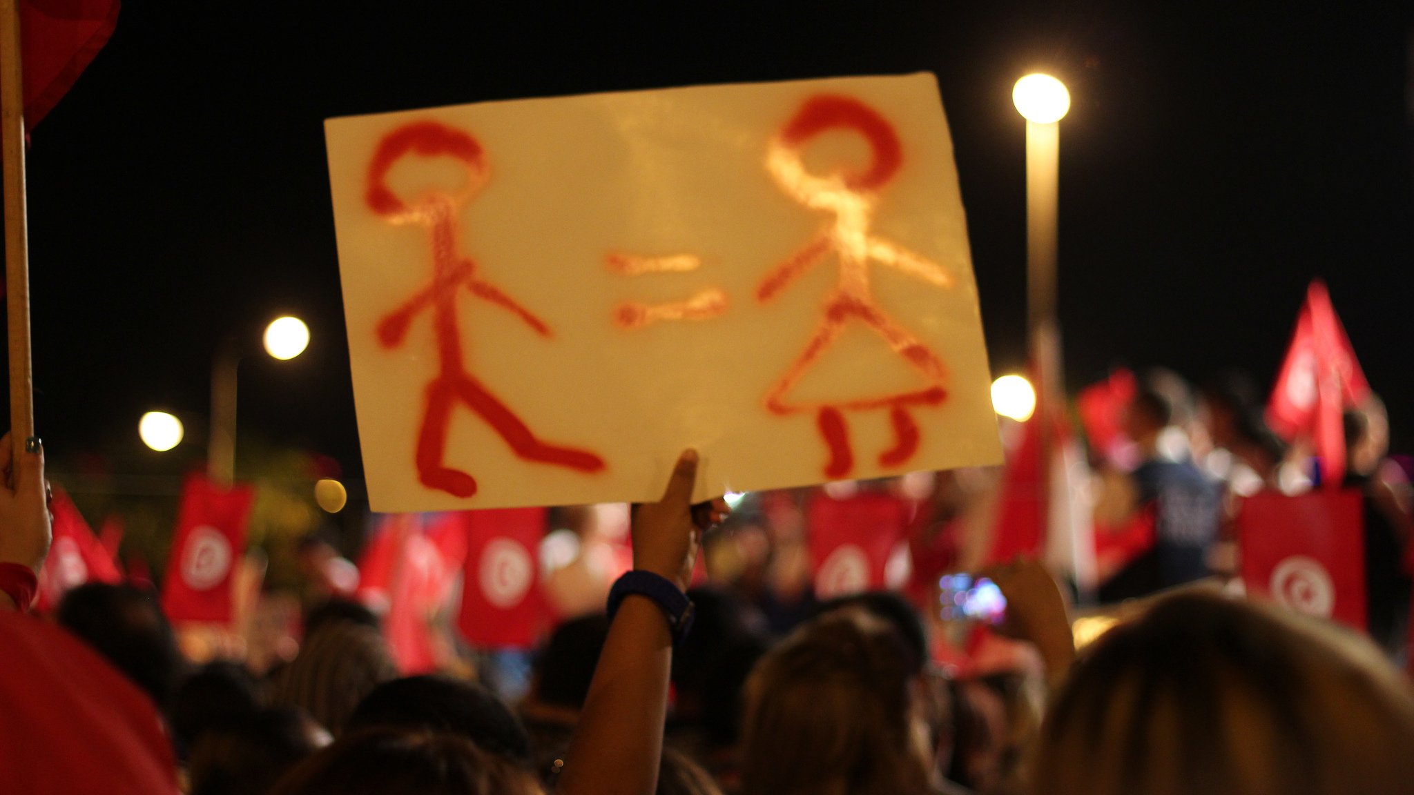 The image shows a night scene of a demonstration or rally, where a person is holding up a sign. The sign features a simplistic representation of a male figure and a female figure, with an equals sign between them, suggesting a message of gender equality. In the background, a crowd can be seen, along with red flags that likely represent a political or social movement. The atmosphere appears to be vibrant and engaged, reflecting a collective message or cause.
