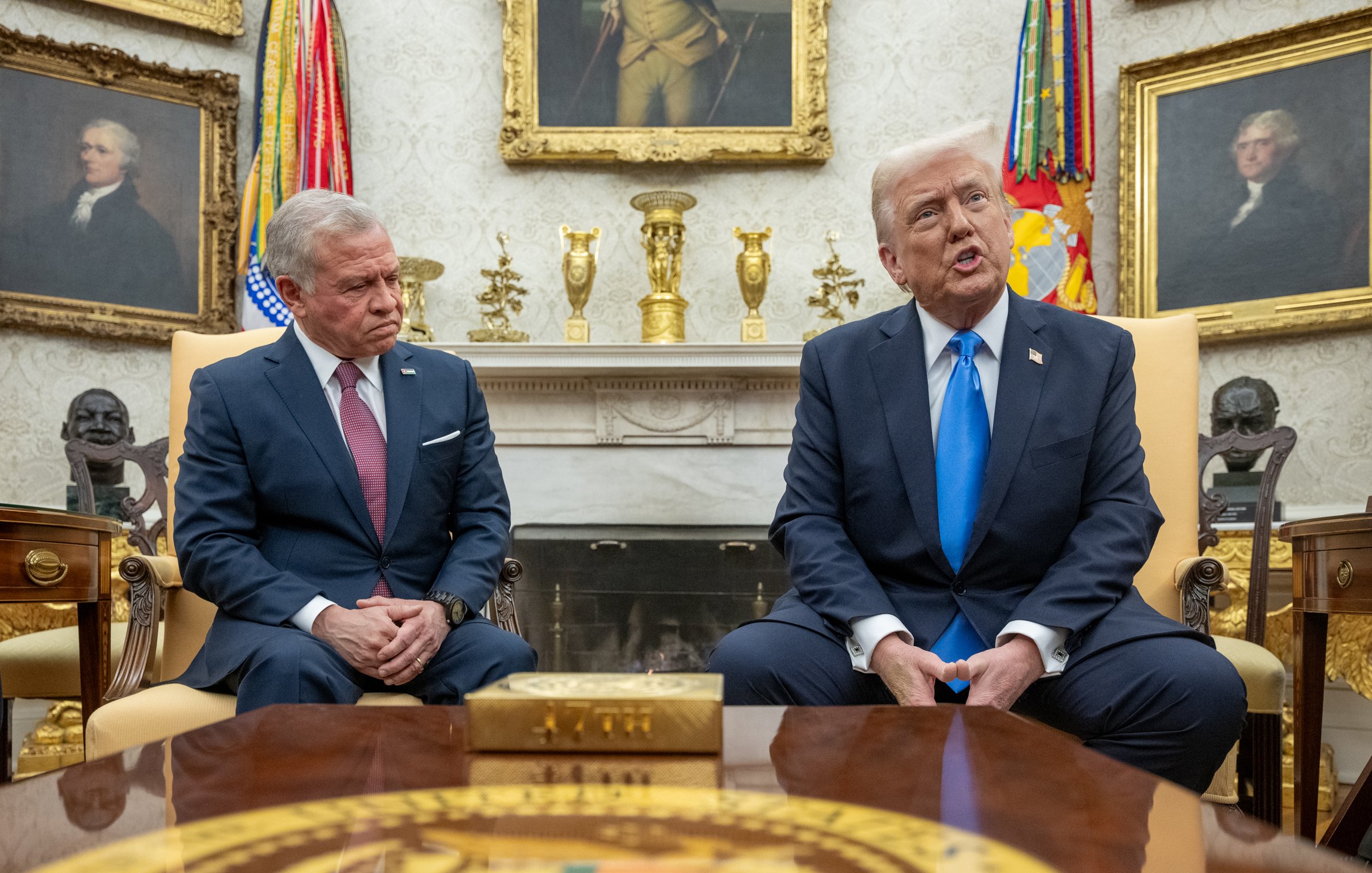 L'image montre deux hommes assis dans une pièce élégante, probablement dans un bureau officiel. L'un d'eux, portant un costume sombre, regarde avec une expression sérieuse, tandis que l'autre, en costume clair avec une cravate bleue, parle. Des drapeaux et des tableaux anciens décorent la pièce. Une table en bois est visible devant eux.
