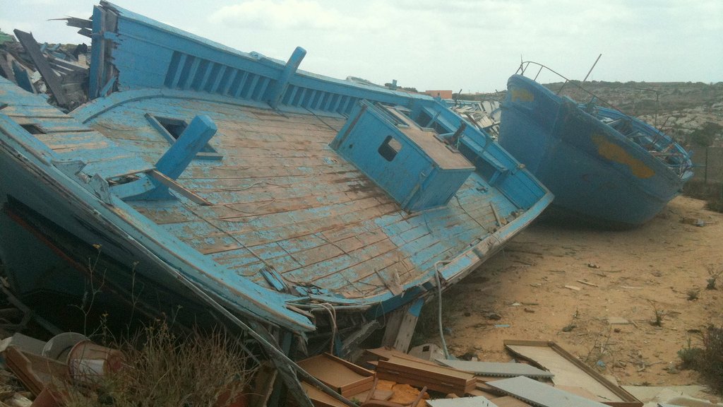 L'image montre des bateaux endommagés, apparemment abandonnés sur une terre aride. Les bateaux, de couleur bleu clair, sont inclinés sur le côté, avec des parties de leur structure exposées. On peut voir des débris et des objets éparpillés autour, ainsi qu'une végétation rudimentaire qui pousse dans le sol. Le ciel est nuageux, créant une atmosphère mélancolique. L'ensemble dégage une impression de désolation et d'abandon.
