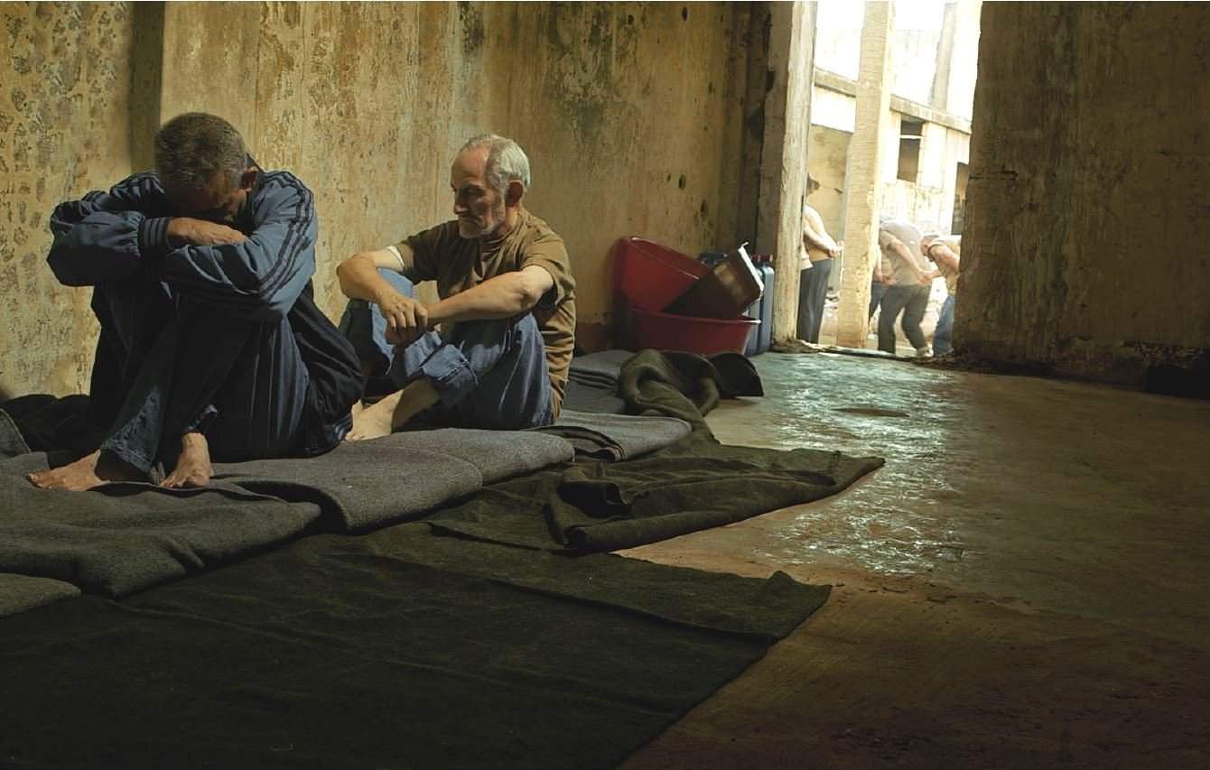 L'image montre un espace intérieur sombre et désolé, probablement une cellule ou un lieu de détention. À l'avant, deux hommes sont assis sur des couvertures posées sur le sol. L'un semble pensif et acculé, tandis que l'autre est plus détendu et semble essayer de communiquer ou de réconforter. En arrière-plan, d'autres personnes sont visibles, marchant en file. L'éclairage est faible, ce qui contribue à une atmosphère de tristesse et de confinement. Les murs semblent usés et abandonnés, renforçant l'impression de désespoir.