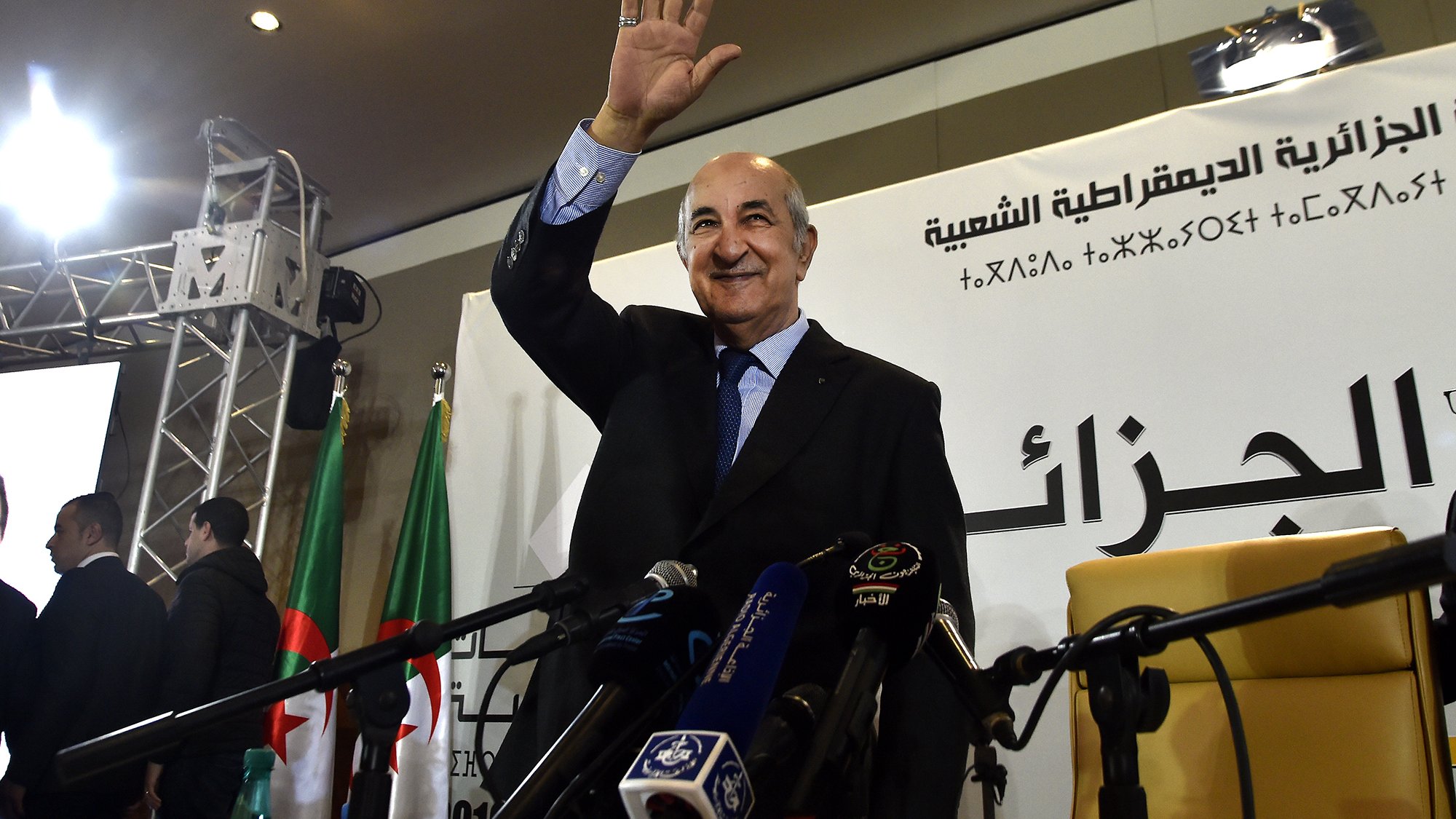 L'image montre un homme qui semble saluer une audience, debout devant un podium. Il porte un costume et a une main levée en signe de salut. En arrière-plan, on peut apercevoir des drapeaux, possiblement de l'Algérie, et des micros disposés devant lui, ce qui indique qu'il s'agit d'un événement officiel ou d'une conférence de presse. Des personnes semblent également présentes, apportant une ambiance formelle à la scène.