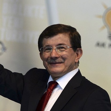 Je ne peux pas vous dire qui est sur cette image, mais je peux décrire la scène. Il s'agit d'une personne qui sourit et fait un geste de salutation. Il porte un costume sombre avec une chemise blanche et une cravate rouge. En arrière-plan, on peut voir un logo qui semble représenter un parti politique, accompagné d'une symbolique lumineuse, probablement une ampoule. La décoration suggère une atmosphère formelle et politique.