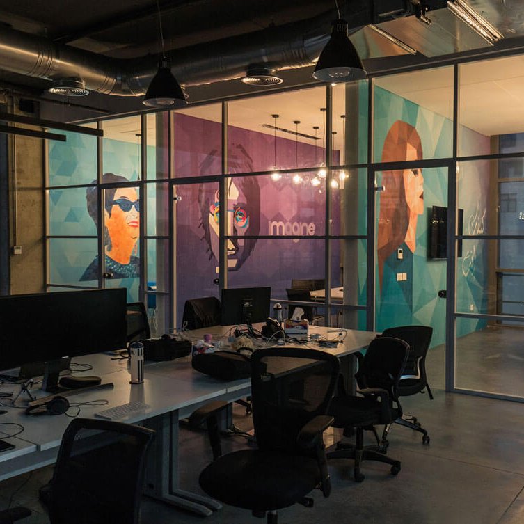 The image shows a modern office space with large glass windows, providing a view of the outside. Inside, there are several workstations with computers and ergonomic chairs. The walls are decorated with vibrant murals featuring stylized portraits of individuals, adding a creative touch to the environment. The overall ambiance is open and contemporary, with industrial elements like exposed ducts and warm lighting fixtures. The space appears to be designed for collaboration and innovation.