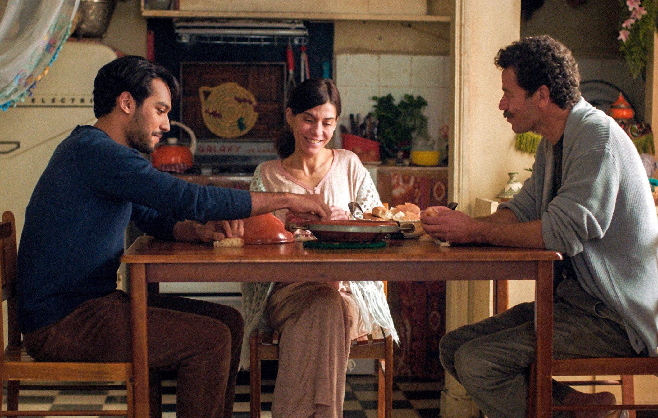 L'image montre trois personnes assises autour d'une table dans une cuisine chaleureuse et accueillante. Il y a un homme à gauche, habillé d'un pull bleu, qui sert de la nourriture à une femme souriante au centre, vêtue d'une blouse claire. À droite, un autre homme, portant un pull gris, semble également engagé dans la conversation. La cuisine est décorée de façon colorée, avec des objets et des ustensiles visibles en arrière-plan, créant une atmosphère conviviale et familiale.