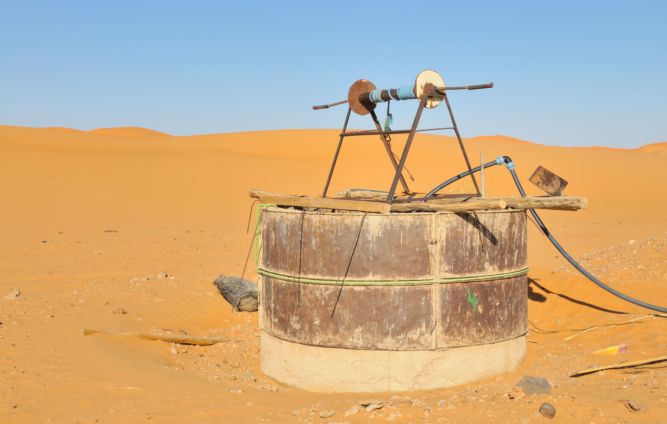 L'image montre une structure située dans un désert de sable doré. Au centre, il y a un puits ou un réservoir en métal, recouvert de tôle, avec un mécanisme en bois au-dessus. Ce dernier semble être utilisé pour pomper de l'eau. La terre environnante est sèche et aride, et on aperçoit des dunes de sable qui s'étendent à perte de vue sous un ciel clair et sans nuages. L'ensemble évoque un environnement désertique, isolé et dépouillé.