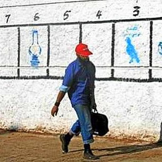 L'image montre un homme marchant le long d'un mur peint en blanc. Sur le mur, il y a une série de numéros de 1 à 8 avec des illustrations ou des symboles correspondant à chaque numéro. L'homme porte une casquette rouge et un vêtement bleu, tenant une mallette. À gauche du mur, une inscription en arabe est visible. L'environnement semble urbain et ensoleillé.