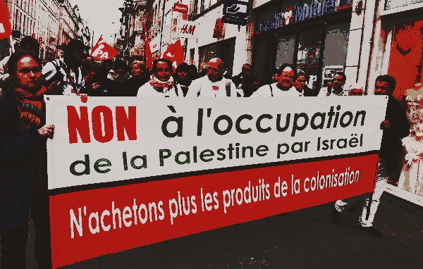 L'image montre une manifestation avec des participants brandissant une grande bannière. Le texte sur la bannière exprime un message de protestation contre l'occupation de la Palestine par Israël, et appelle à ne plus acheter les produits de la colonisation. Les manifestants semblent unis et sont vêtus de vêtements variés, avec des drapeaux en arrière-plan. L'ambiance est sérieuse et engagée, reflétant un message politique fort.