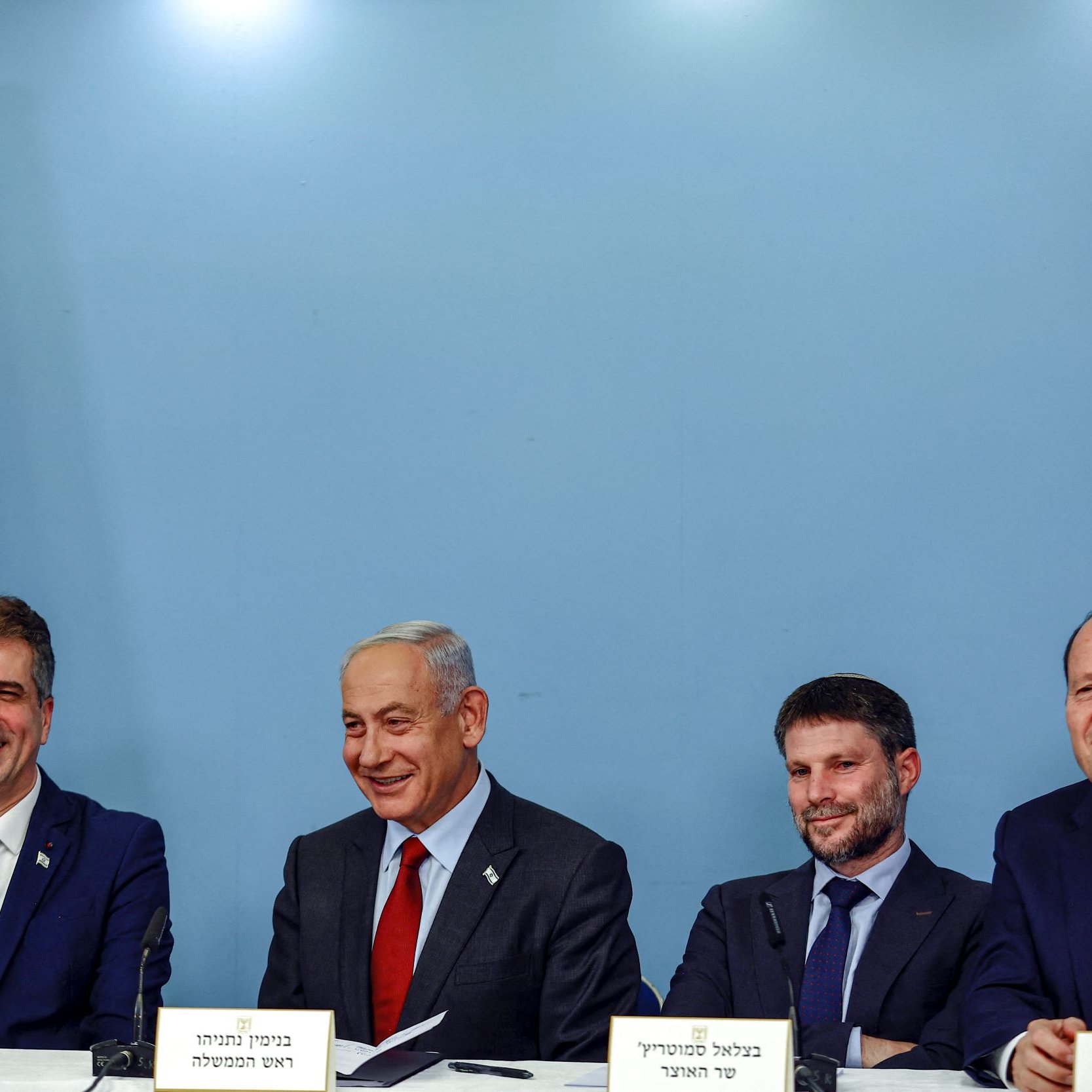 The image shows a group of four men seated at a table in front of a blue backdrop with the Israeli flag. They appear to be engaged in a discussion or announcement. The man in the center, likely the most prominent figure, is smiling. Each individual has a nameplate in front of them, indicating their identities or titles. The overall atmosphere seems formal, suggesting a political or governmental context.