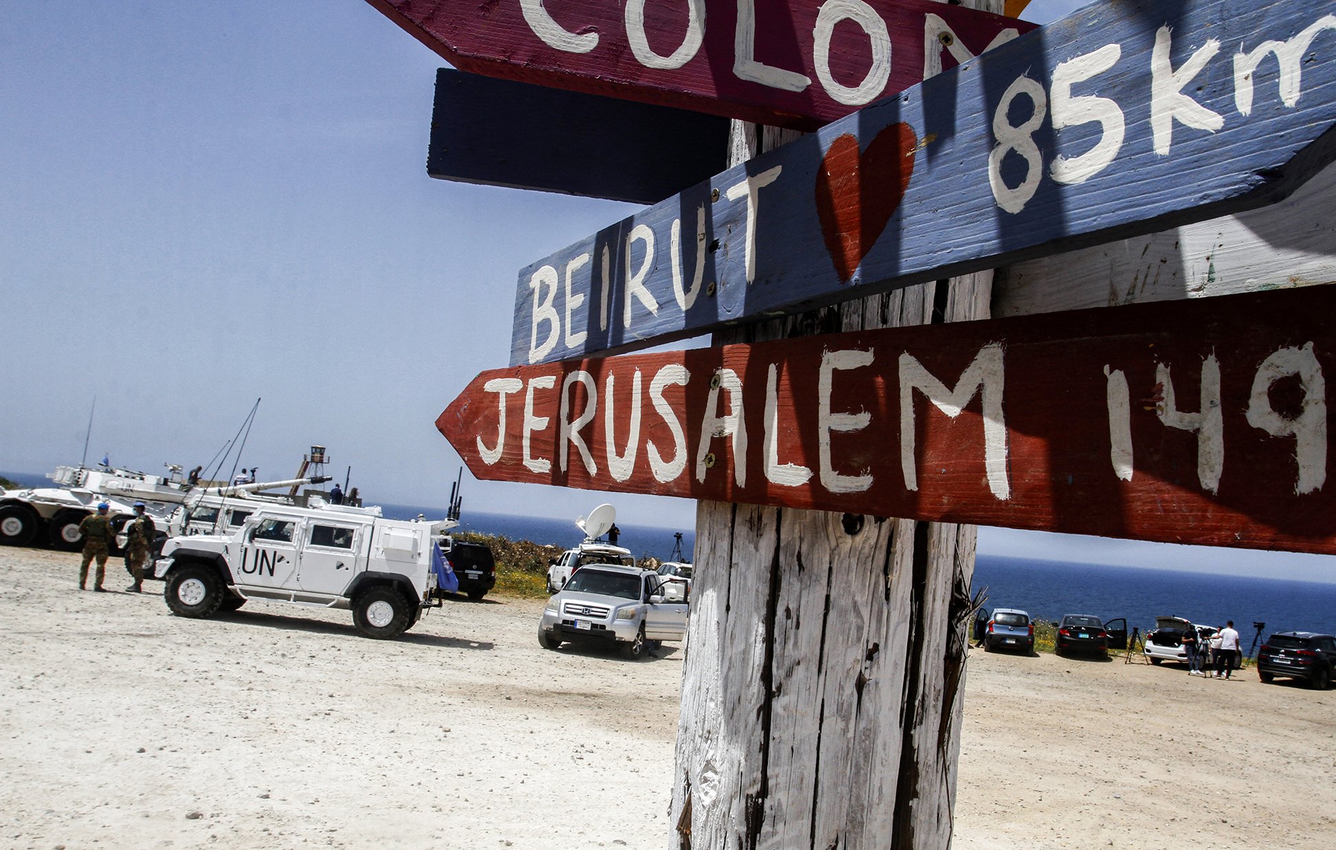 The image depicts a wooden signpost with several directional arrows pointing towards different locations. The names of the places, including "Beirut" and "Jerusalem," are painted in bold, colorful letters. The distances to these locations are also indicated in kilometers. In the background, there are vehicles, including a UN truck, parked on a sandy area near the coastline, with a view of the sea. The overall setting appears to be a scenic lookout or border area.