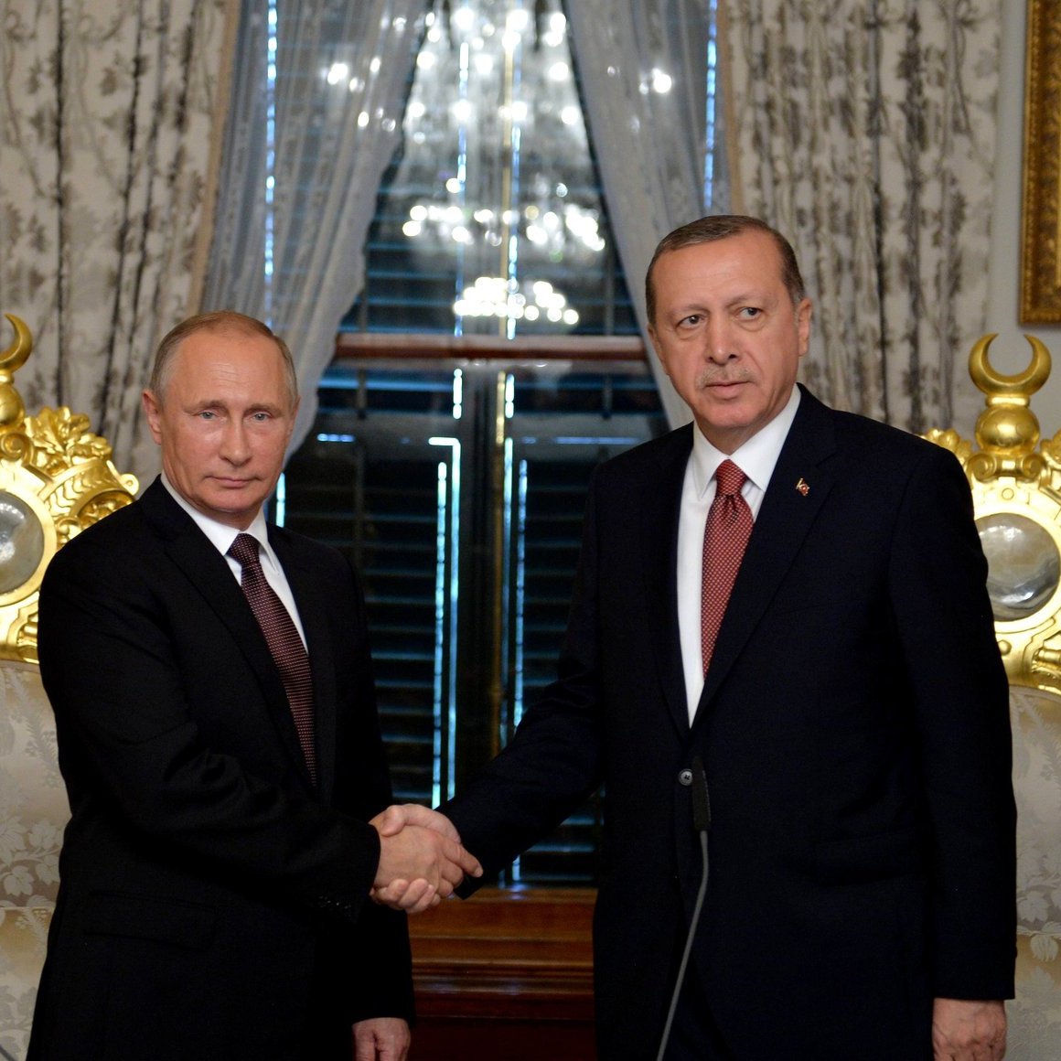 تصویر نشان‌دهنده‌ی دو نفر است که در حال握 دستی هستند. آن‌ها در یک محیط رسمی قرار دارند و در پس‌زمینه، دکوراسیون مجلل و البته دو پرچم، یکی از روسیه و دیگری از ترکیه، دیده می‌شود. این صحنه می‌تواند به یک ملاقات یا اجلاس رسمی بین مقامات دو کشور اشاره داشته باشد. فضای تصویر به طور کلی جدی و رسمی به نظر می‌رسد.