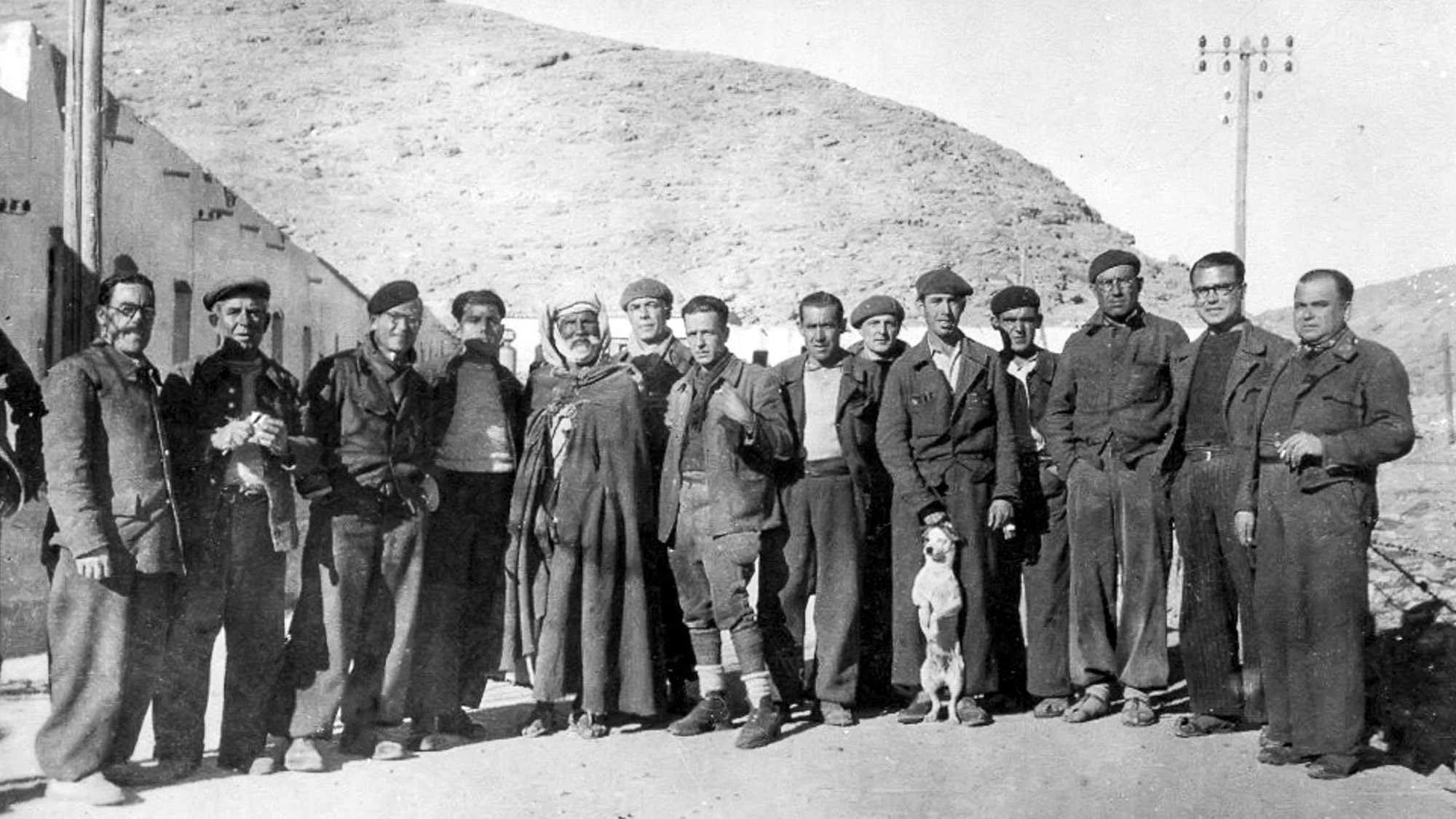 L'image en noir et blanc montre un groupe d'hommes se tenant debout ensemble, probablement dans un environnement rural ou montagneux. Ils portent des vêtements qui semblent être des tenues de travail ou militaires. Au centre, un homme en costume traditionnel se distingue des autres. Un chien se trouve également parmi le groupe. Les hommes affichent diverses expressions, allant des sourires aux regards sérieux. L'arrière-plan présente des collines, et quelques bâtiments peuvent être aperçus. L'ensemble évoque une période historique, probablement au début ou au milieu du 20e siècle.