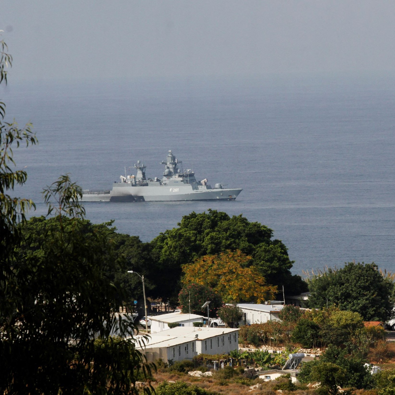 L'image montre un navire militaire se trouvant dans une étendue d'eau, probablement en mer. On aperçoit des paysages côtiers avec quelques arbres et des bâtiments en arrière-plan. Le navire a une structure moderne et est visible au loin, tandis que la végétation au premier plan donne une impression de profondeur à la scène. L'atmosphère semble claire, suggérant une journée ensoleillée.