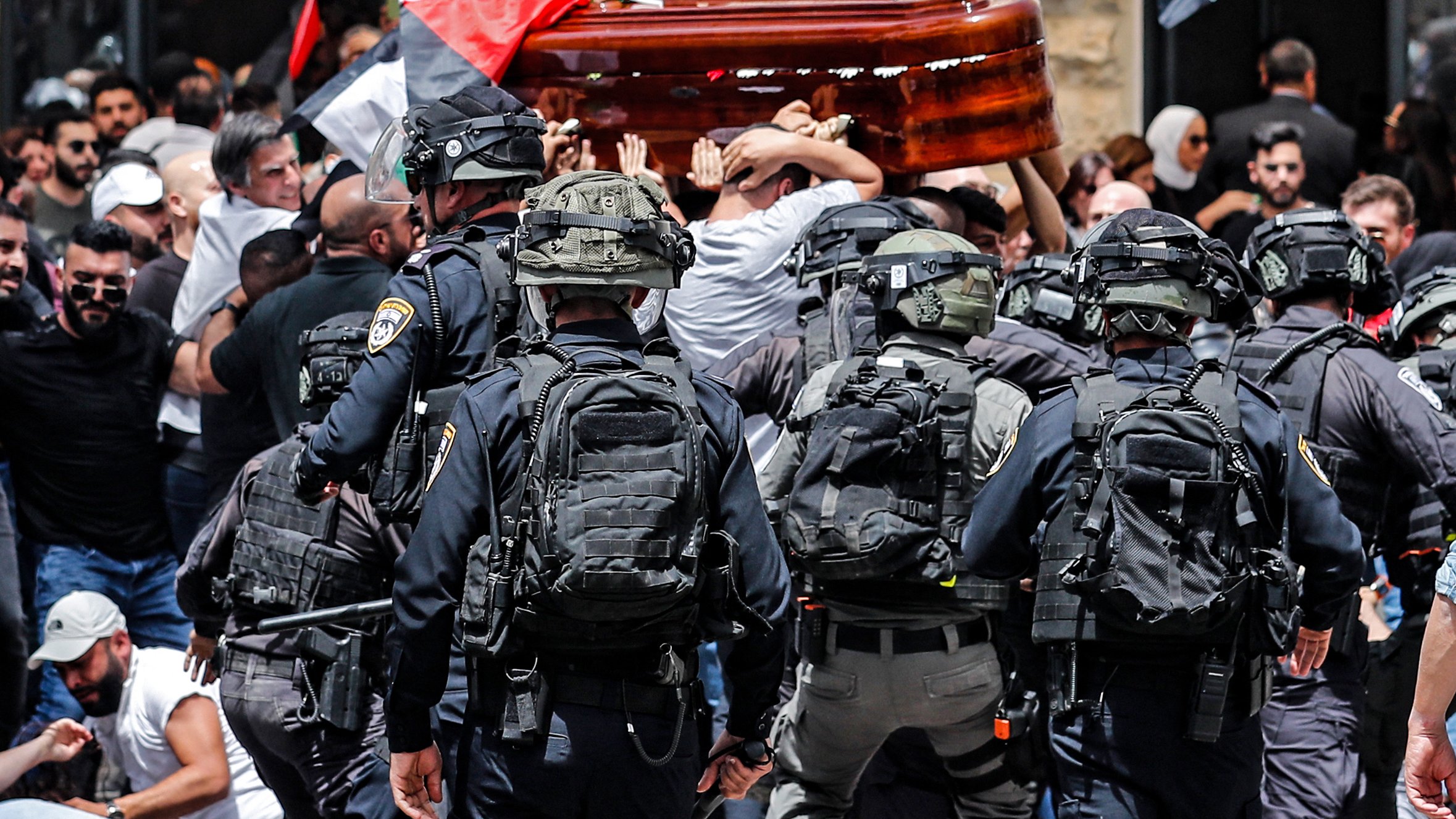 L'immagine rappresenta una scena di tensione durante una manifestazione. Ci sono persone che trasportano un barella, probabilmente durante un funerale o una commemorazione, mentre un gruppo di agenti di polizia in equipaggiamento anti-sommossa sta cercando di mantenere l'ordine. Molti manifestanti sono visibili, con bandiere che simboleggiano le loro rivendicazioni, e l'atmosfera appare carica di emozione e conflitto. La scena è caratterizzata da un forte senso di mobilitazione e protesta.